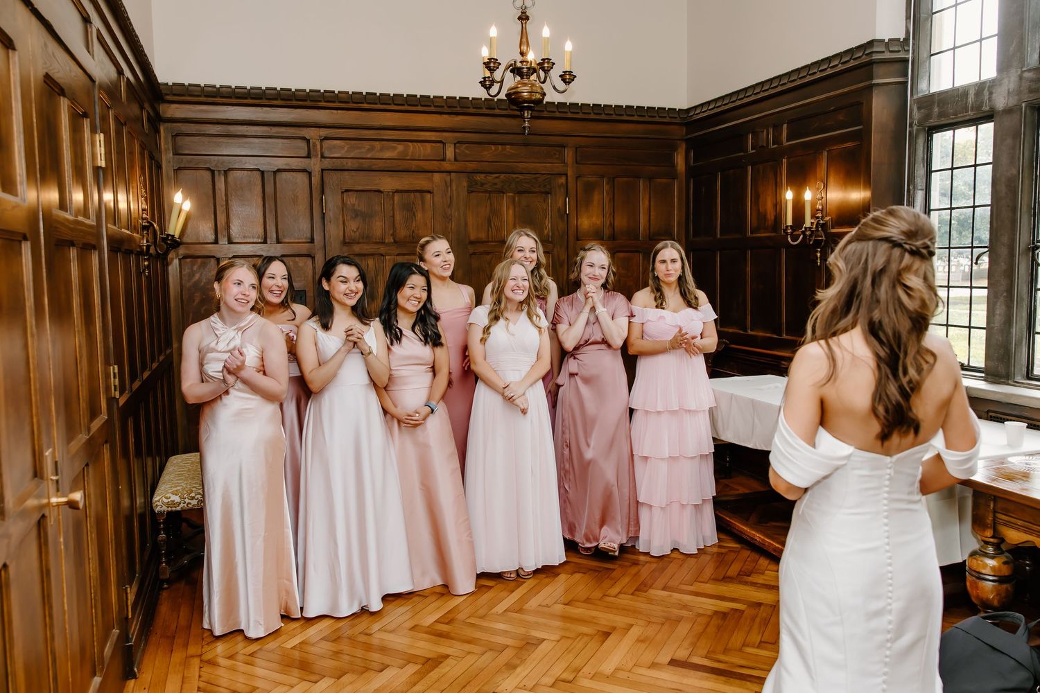 Bridesmaids in pink dresses stand together in a grand wood-paneled room with chandelier.