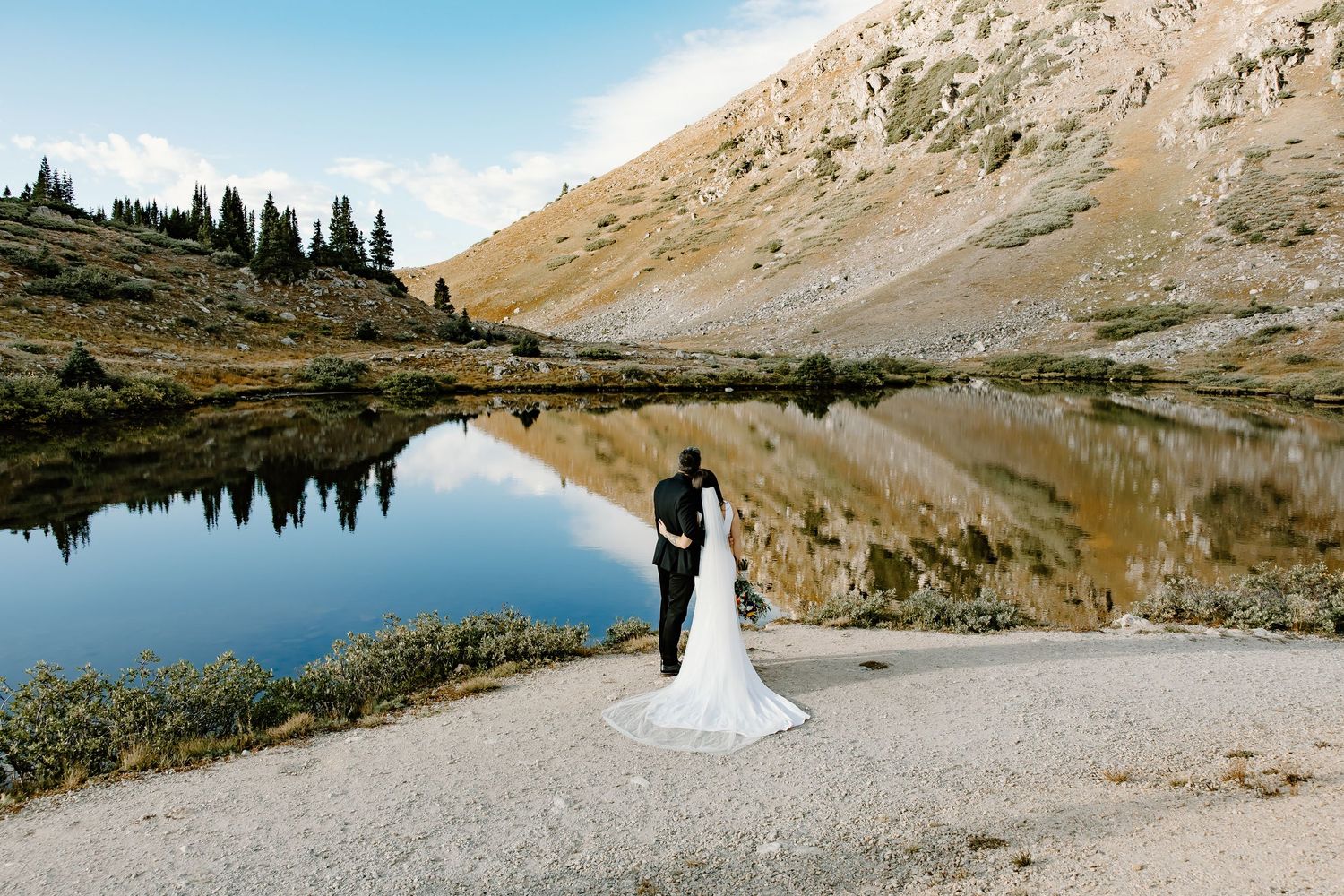 Panoramic mountain lake view with a couple celebrating their wedding on the rocky shore.