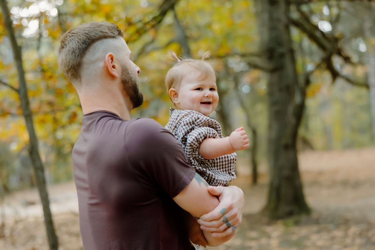 Parent lifts happy child in checkered outfit against autumn forest backdrop.