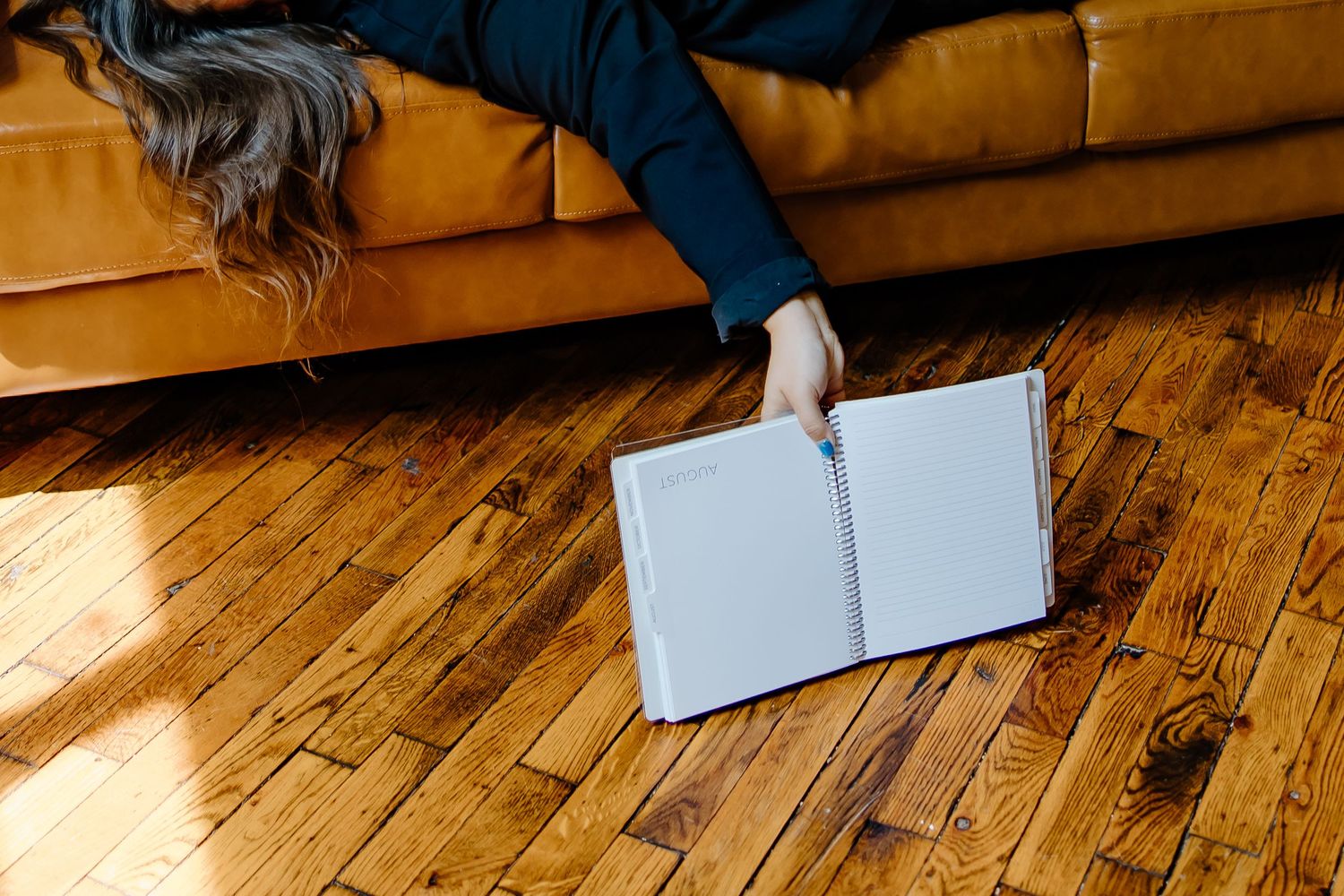 Hand reaching down to pick up a white book or album from a sunlit hardwood floor next to an orange velvet couch.