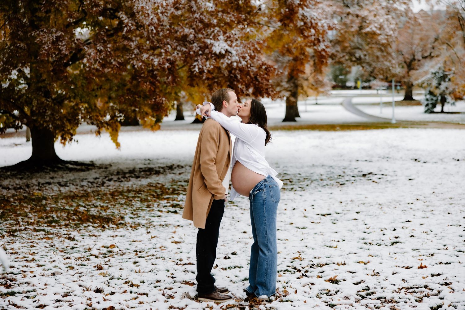 Expectant couple embracing on snowy ground with autumn trees in background.