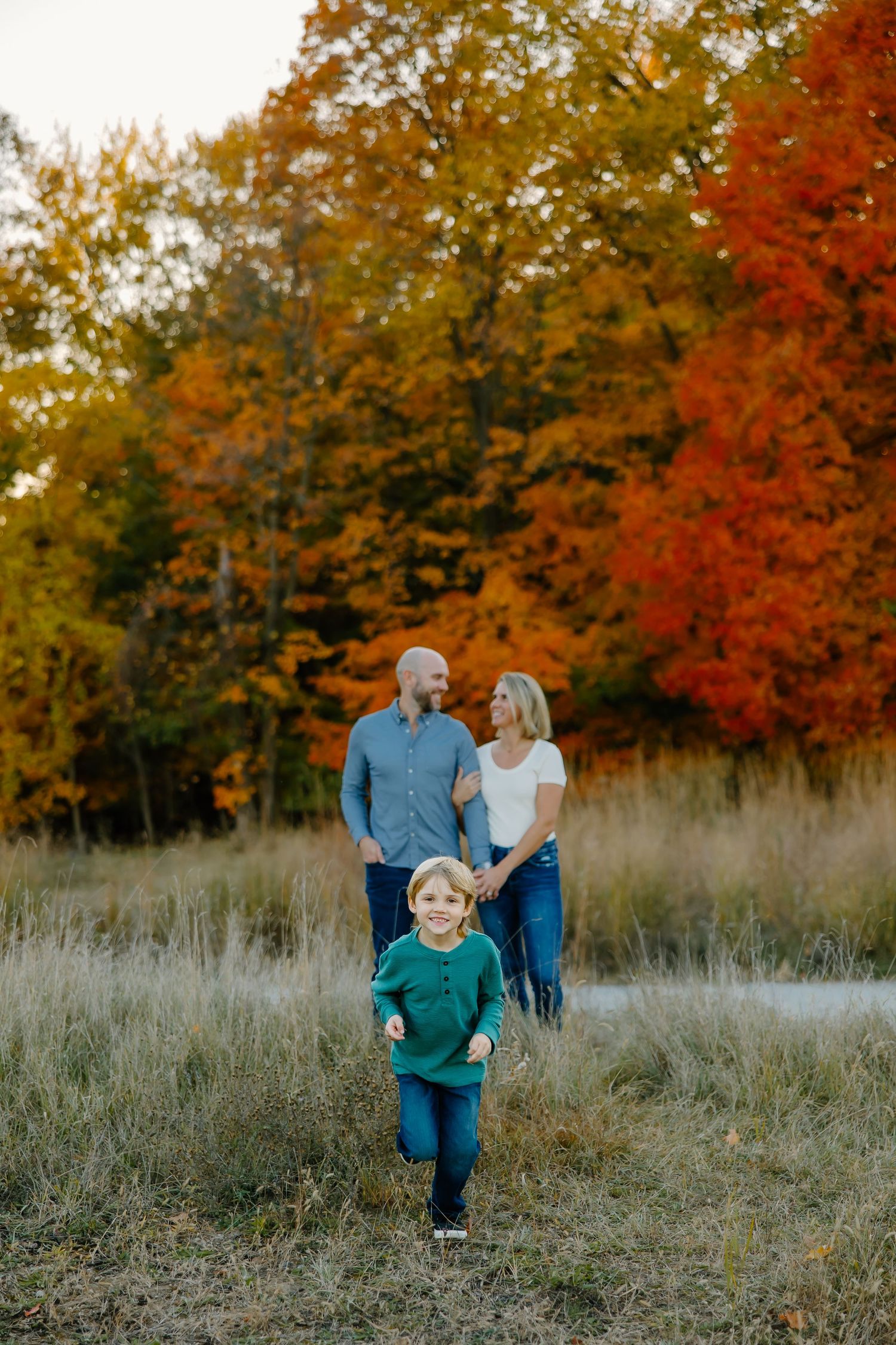 A family stands together in a grassy field during autumn with vibrant orange and yellow foliage in the background.