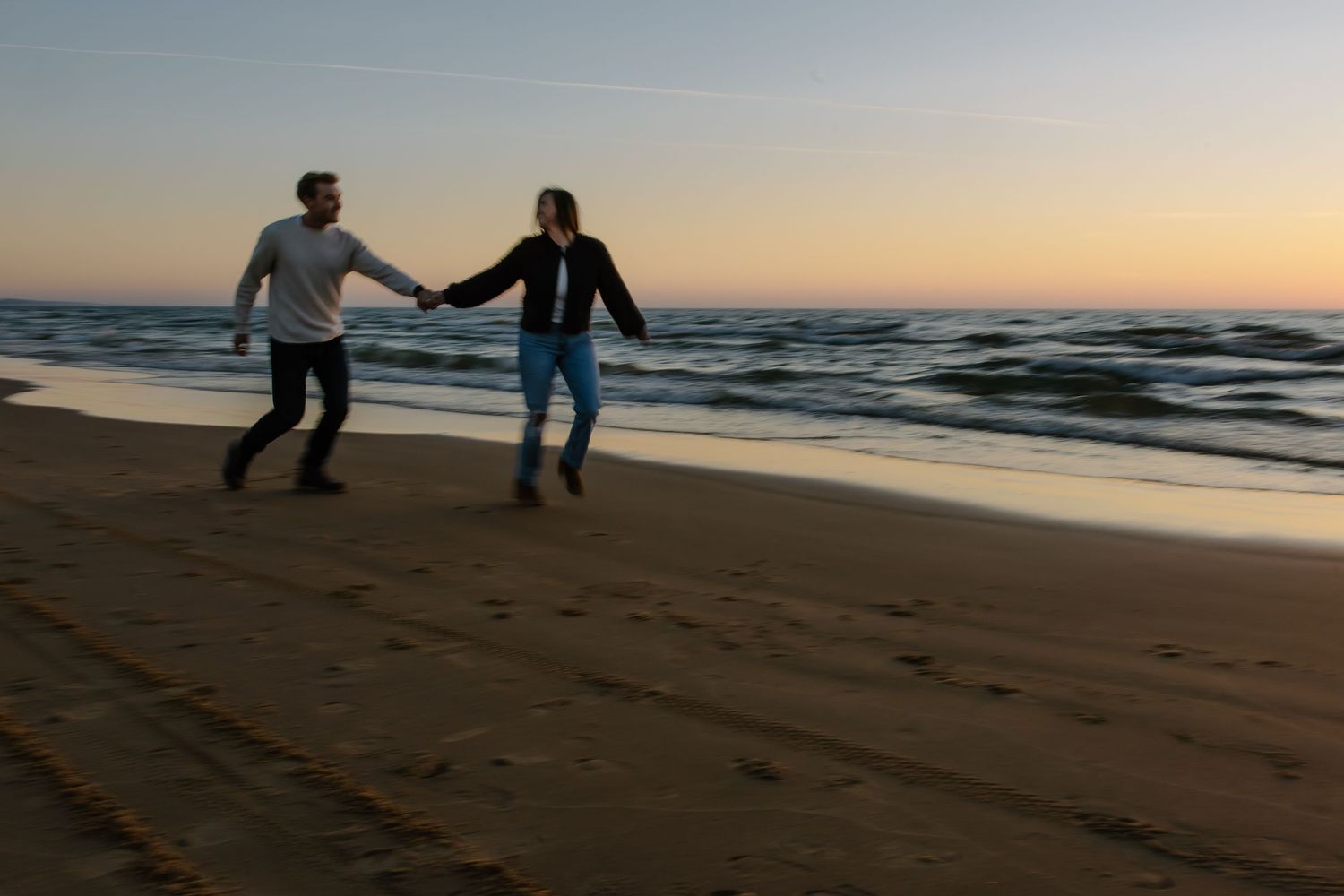 Couple playfully dancing together on a beach at sunset with ocean waves in the background.