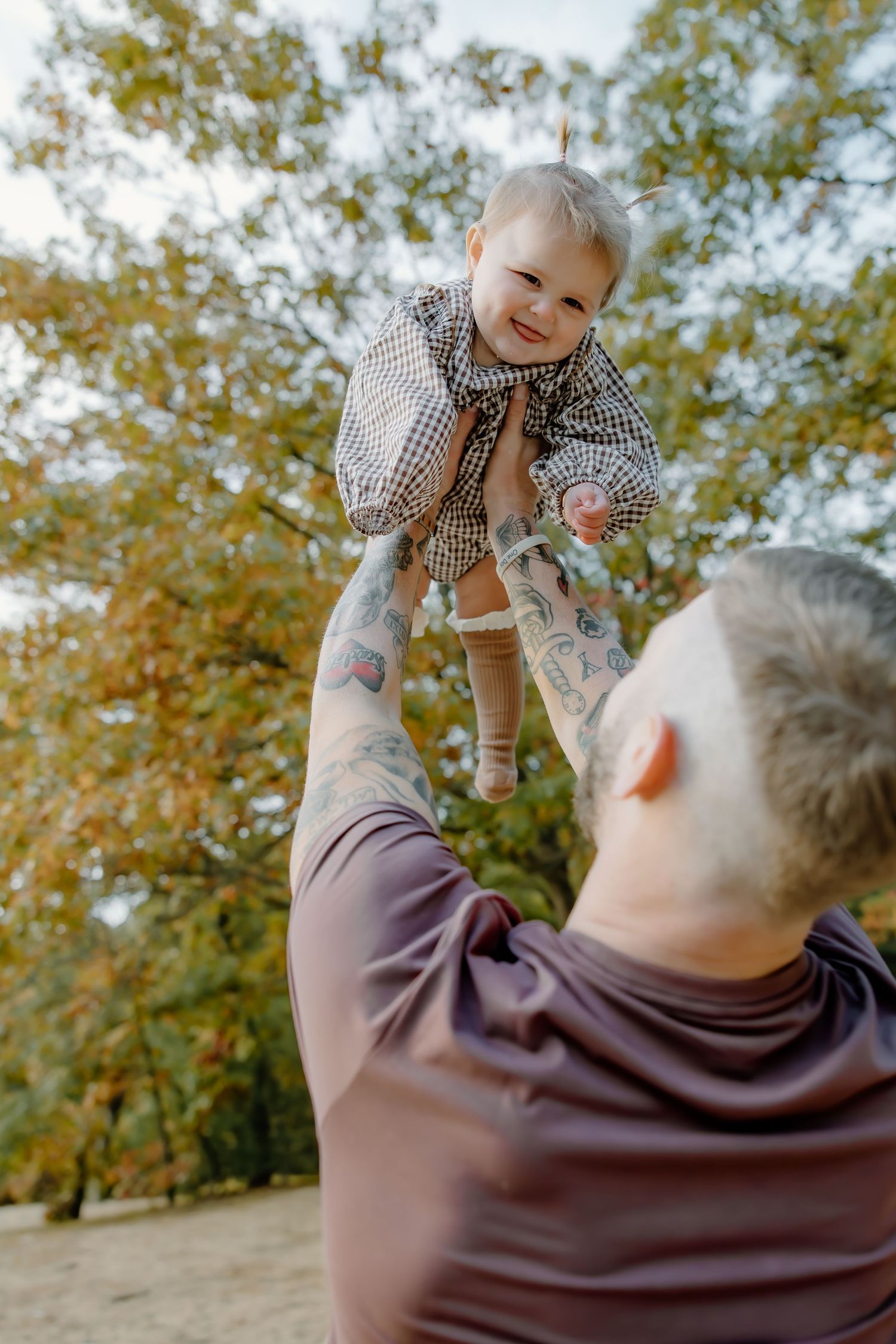 Person in gray sweater lifts small child wearing checkered outfit against autumn trees.