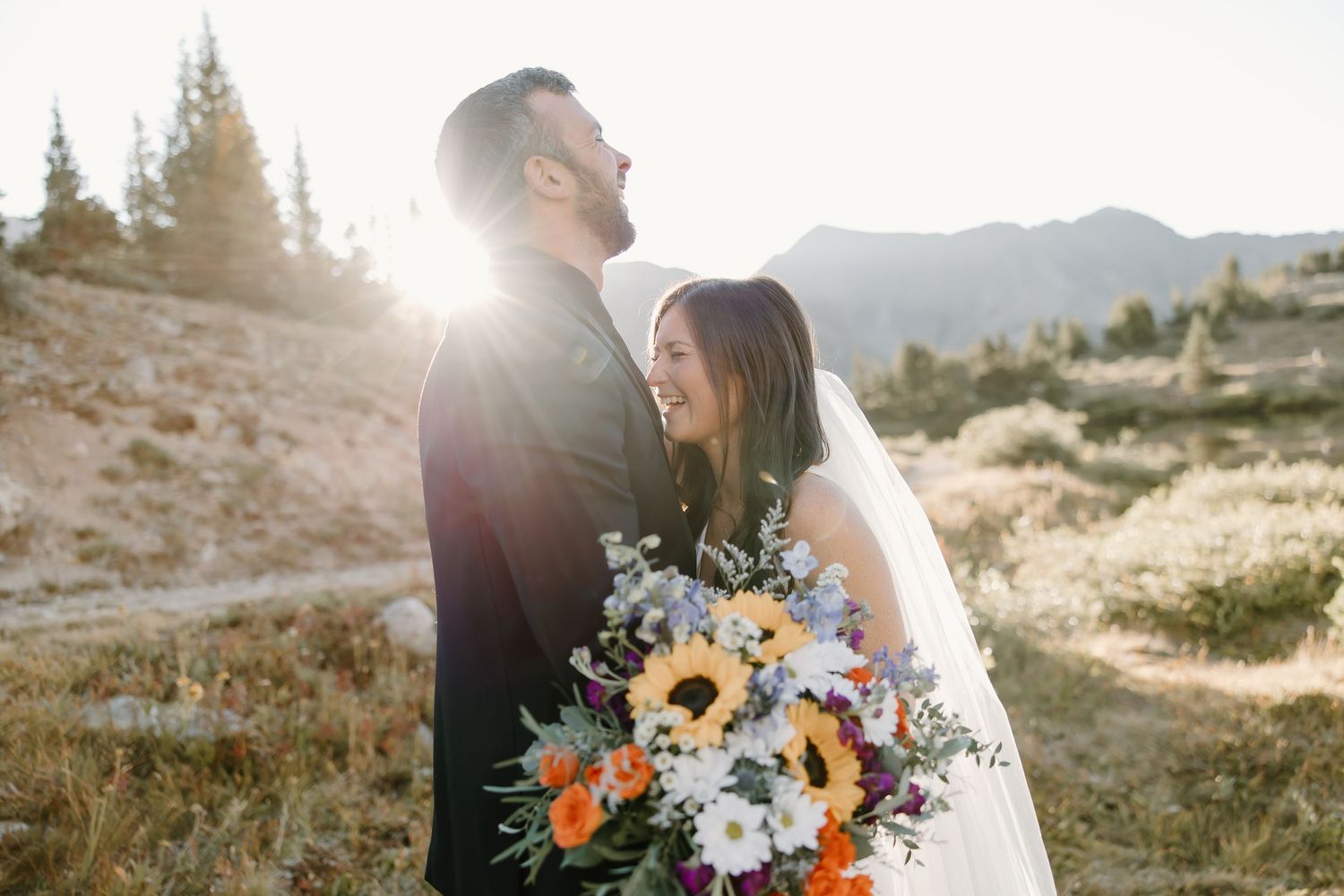Newlyweds embrace in a mountain meadow during sunset with a colorful wildflower bouquet.