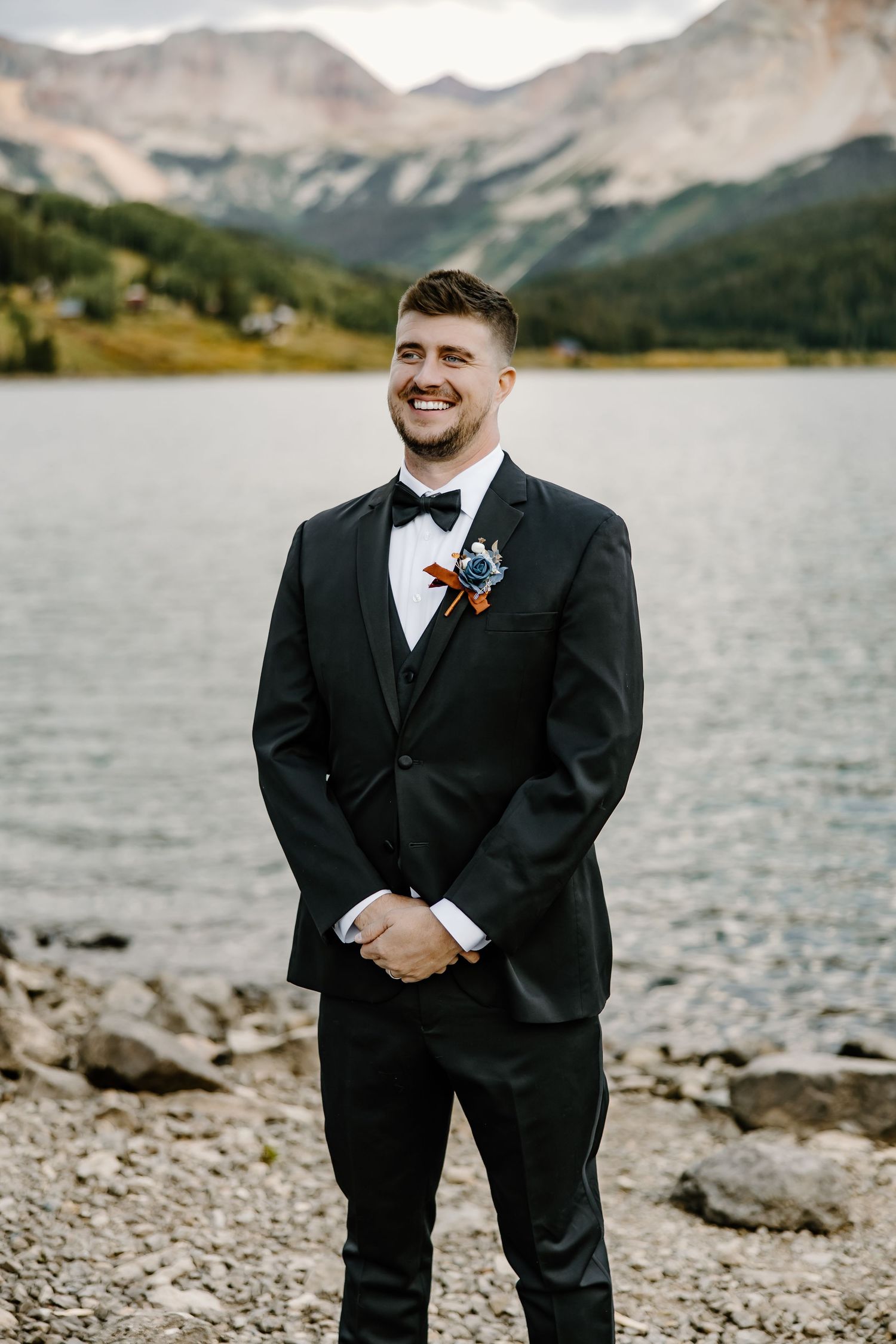 Person in formal black suit standing by mountain lake with rocky shore.
