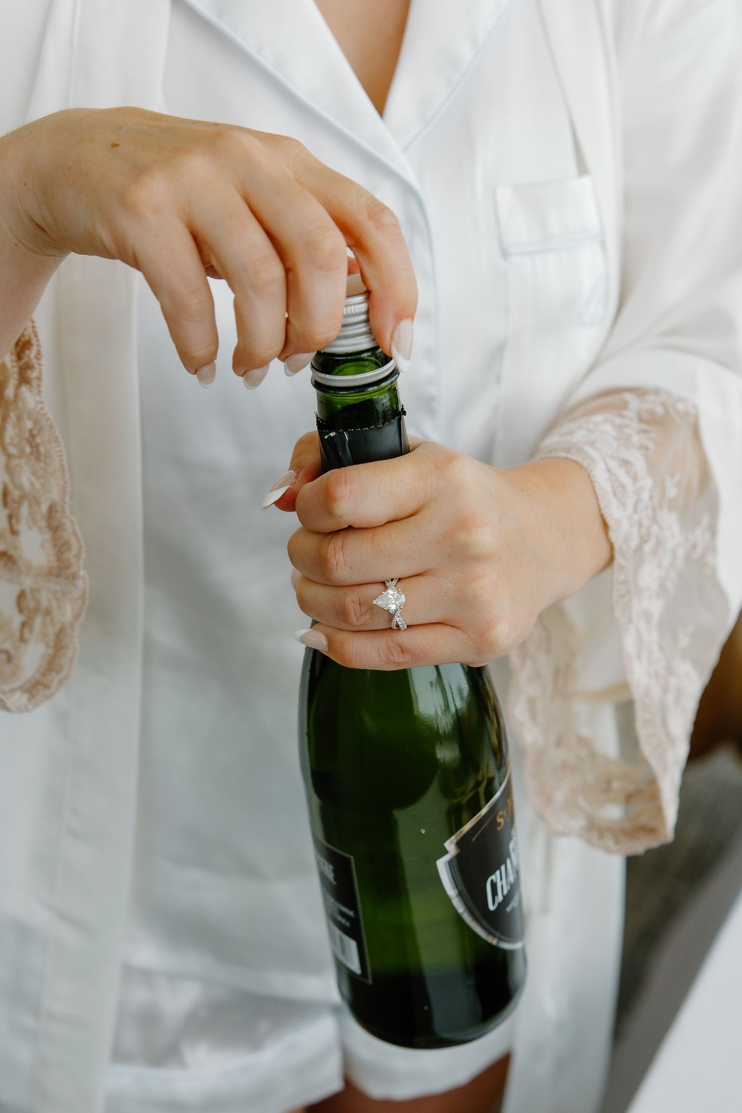 Hands wearing an engagement ring opening a champagne bottle while wearing white lace robe.