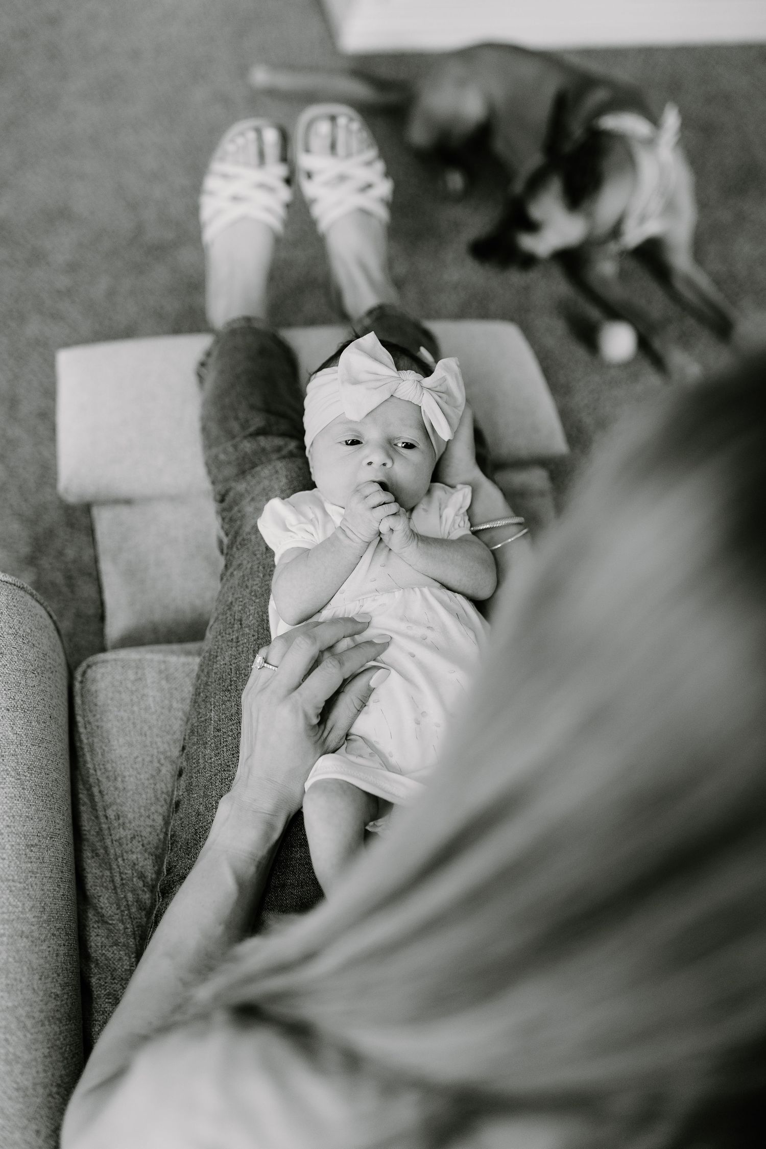 Black and white photo of a small baby in white clothing resting on a couch with a dog visible in the background.