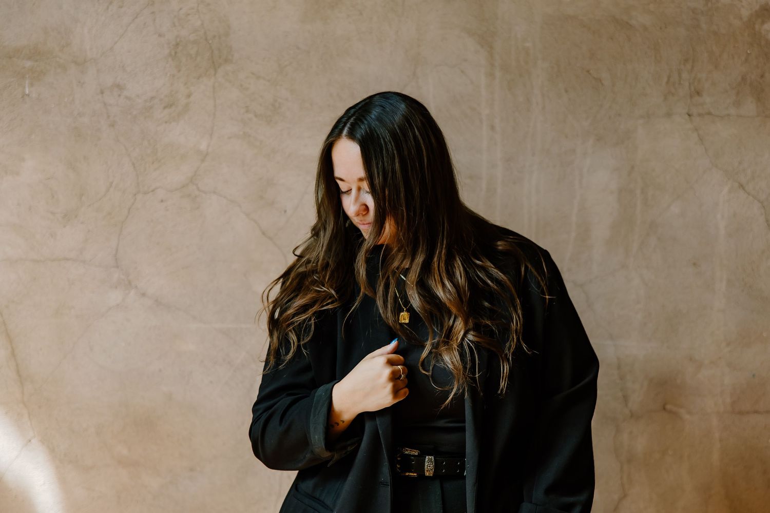 A person wearing a black coat stands against a textured beige wall in a moody artistic portrait series.