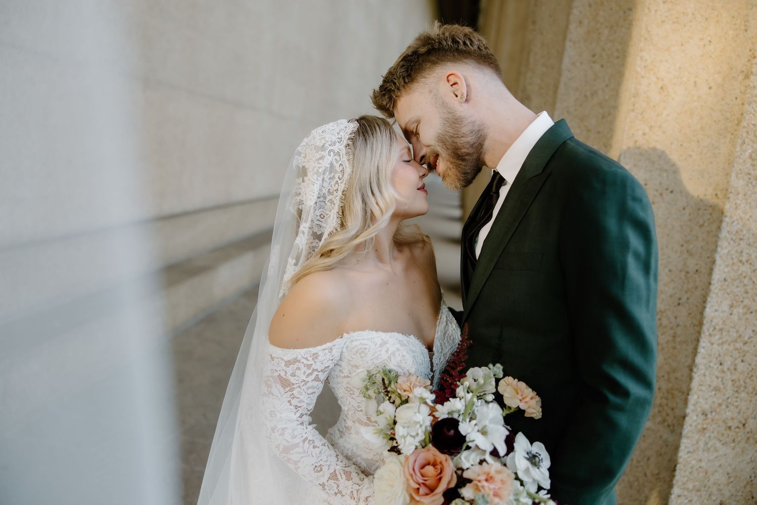 Bride and groom share romantic moment, touching foreheads, bride in lace gown with veil holding floral bouquet.