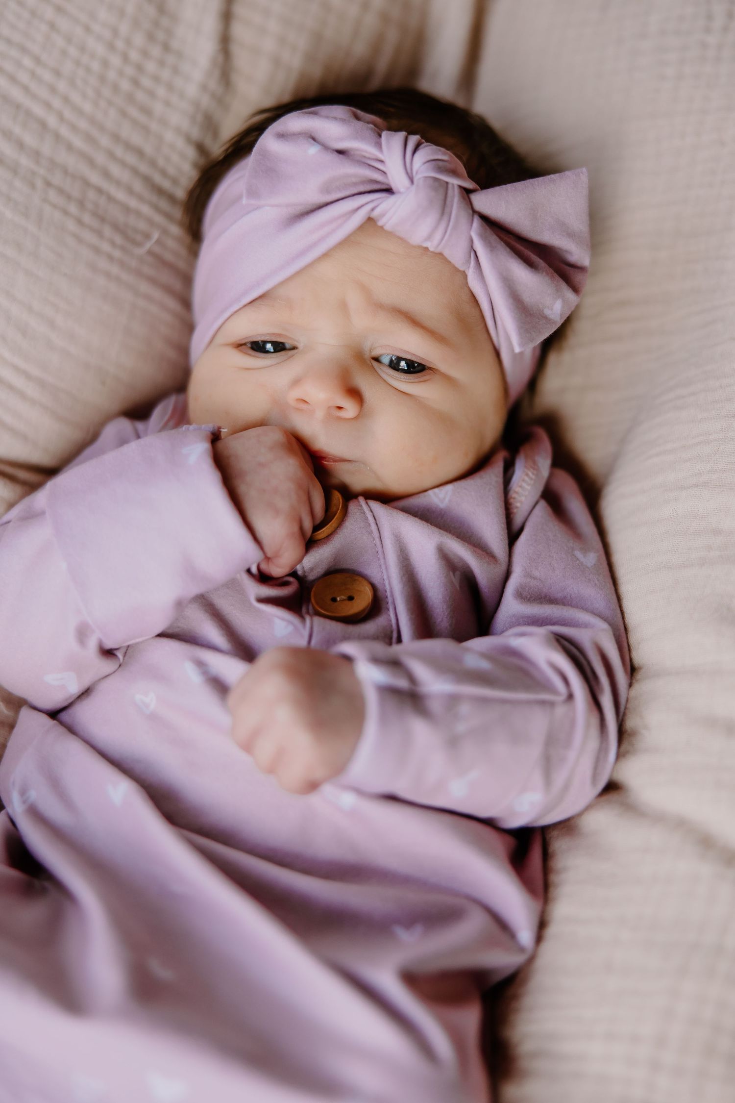 A baby wearing a pink outfit and matching bow headband lies on soft cushions.