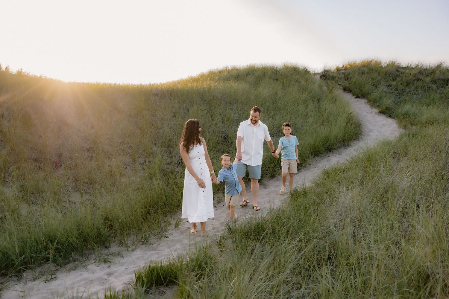 Family of four walking on a sandy path through green dunes at golden hour sunset.