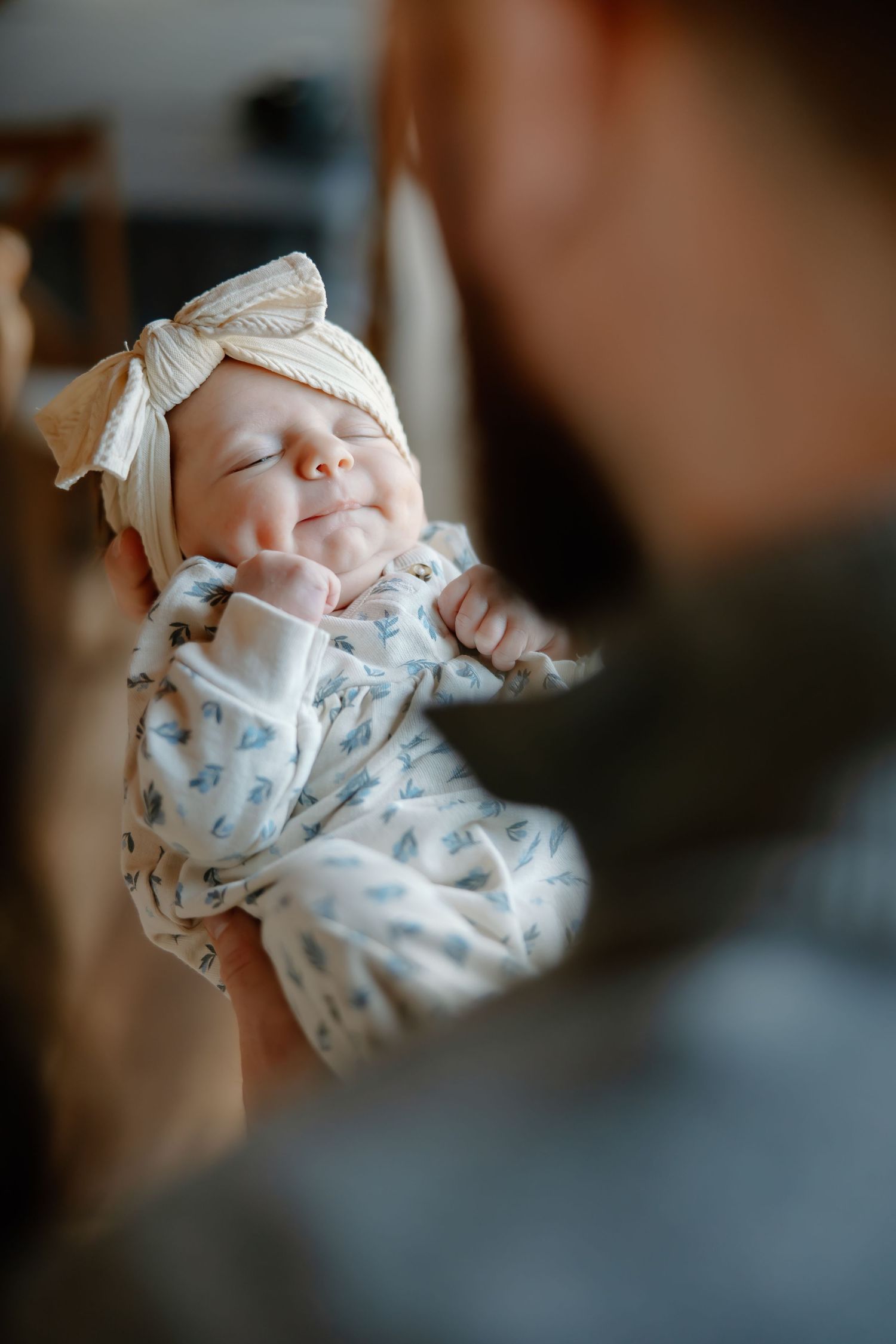 A sleeping newborn baby wearing a bow headband and floral onesie smiles contentedly.