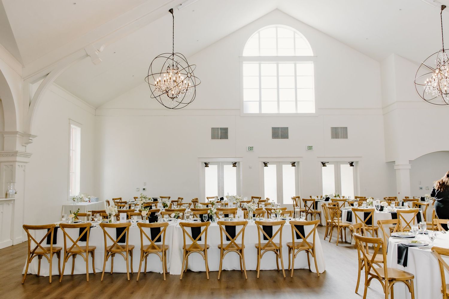 Long banquet tables with wooden chairs set up in an all-white wedding reception hall with chandeliers.
