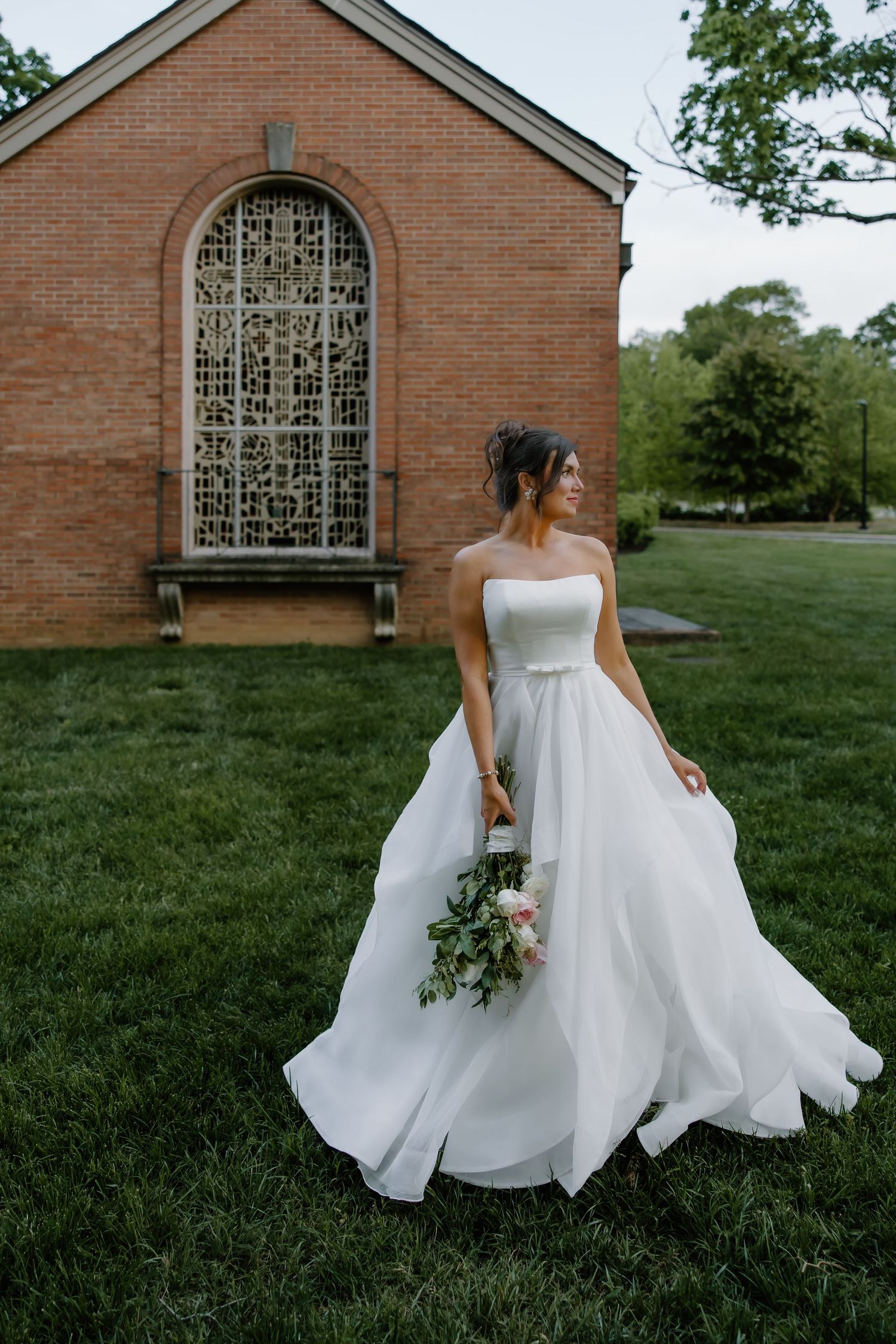 Bride in strapless ballgown poses in front of brick chapel with ornate window.