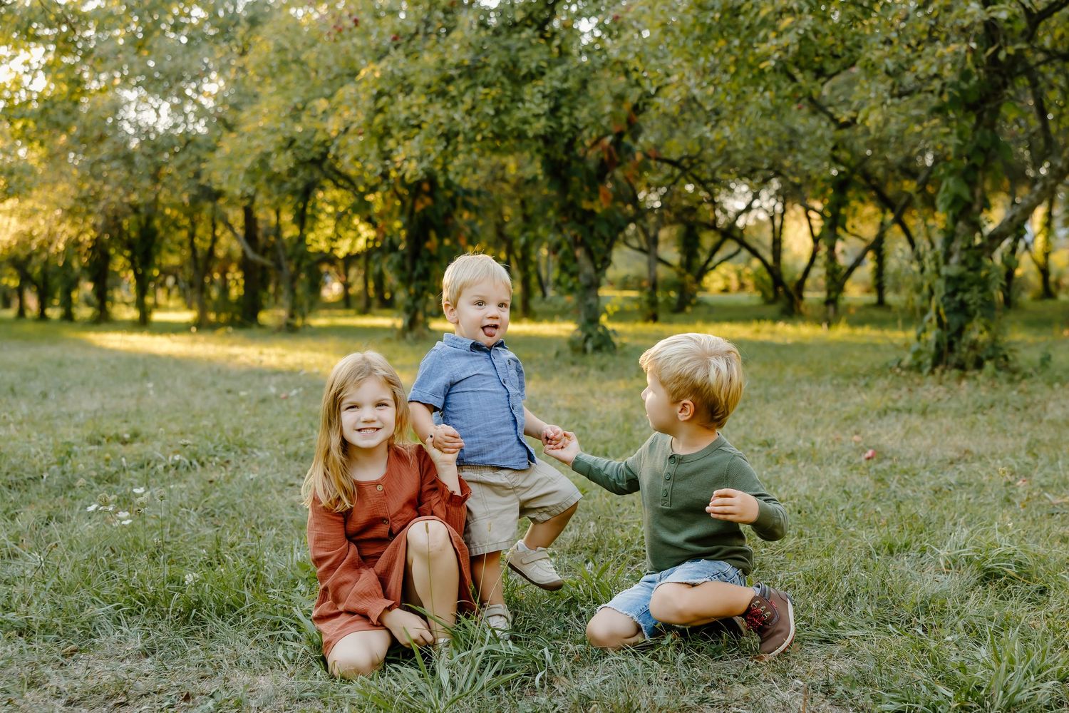 Three children playing together on grass in an autumn orchard setting.