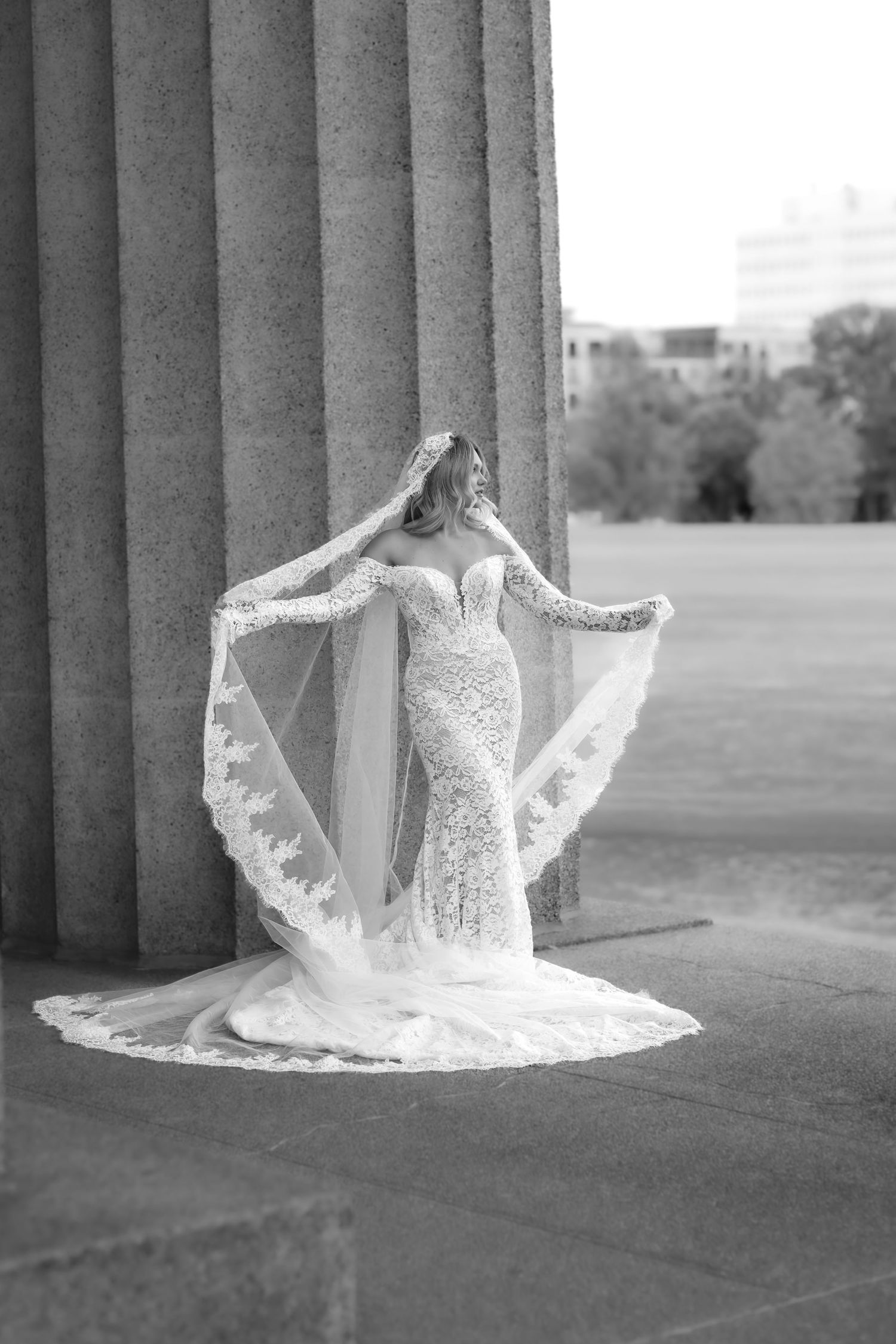 Bride in lace mermaid gown with flowing veil poses between stone columns in black and white bridal portrait.