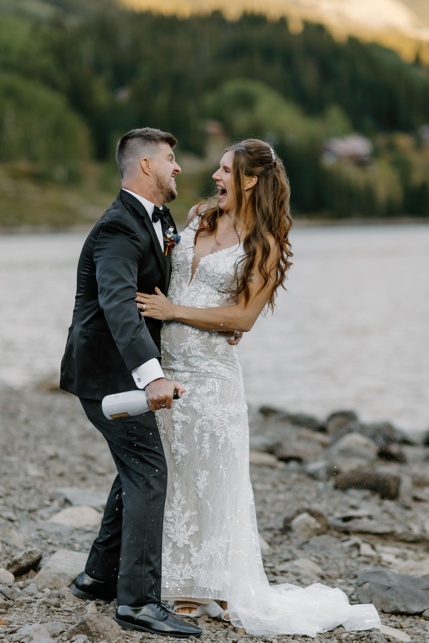A joyful wedding couple shares a laugh on a rocky lakeshore with mountains in the background.