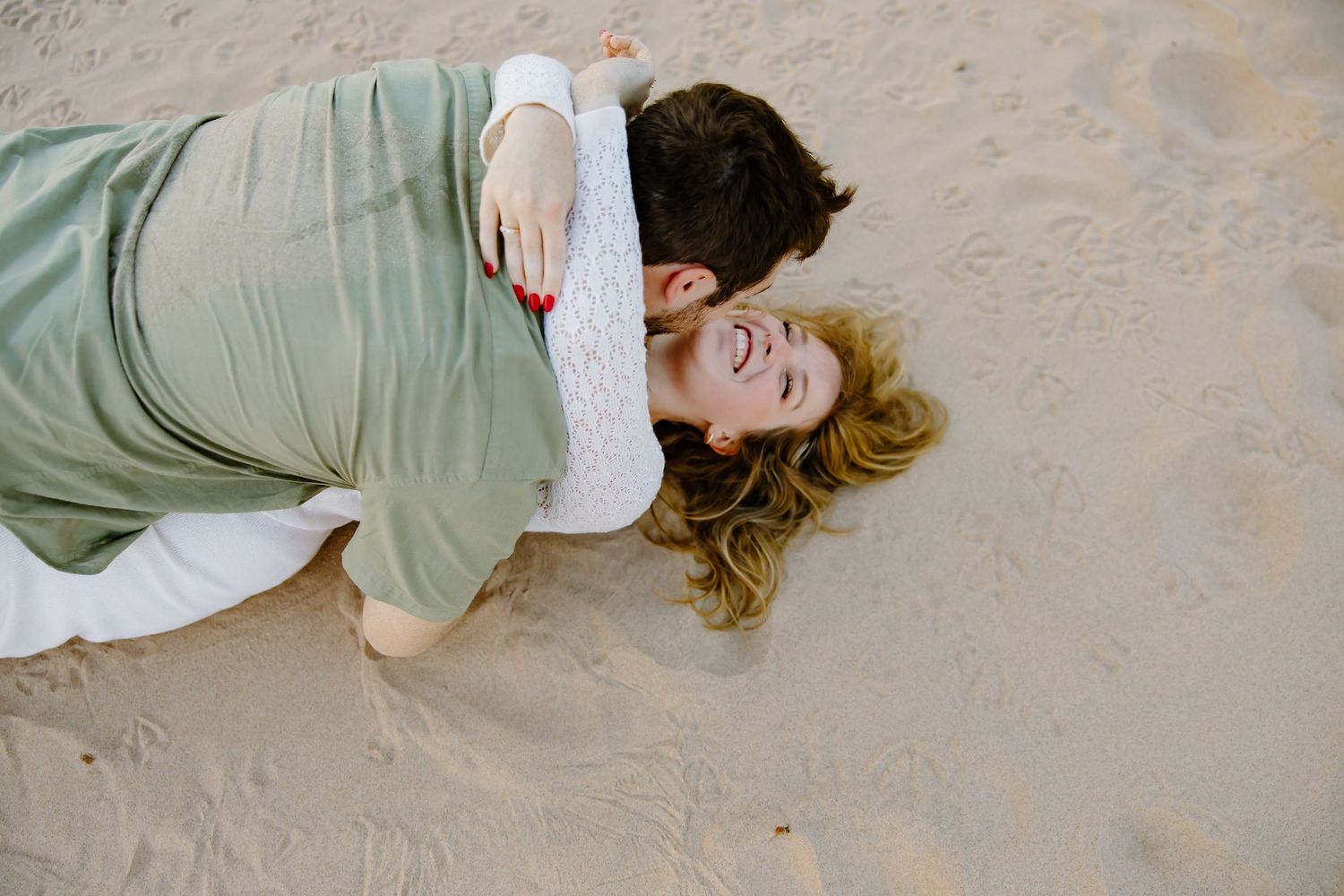 A playful moment captured as a couple lies in the sand during their beach photoshoot.
