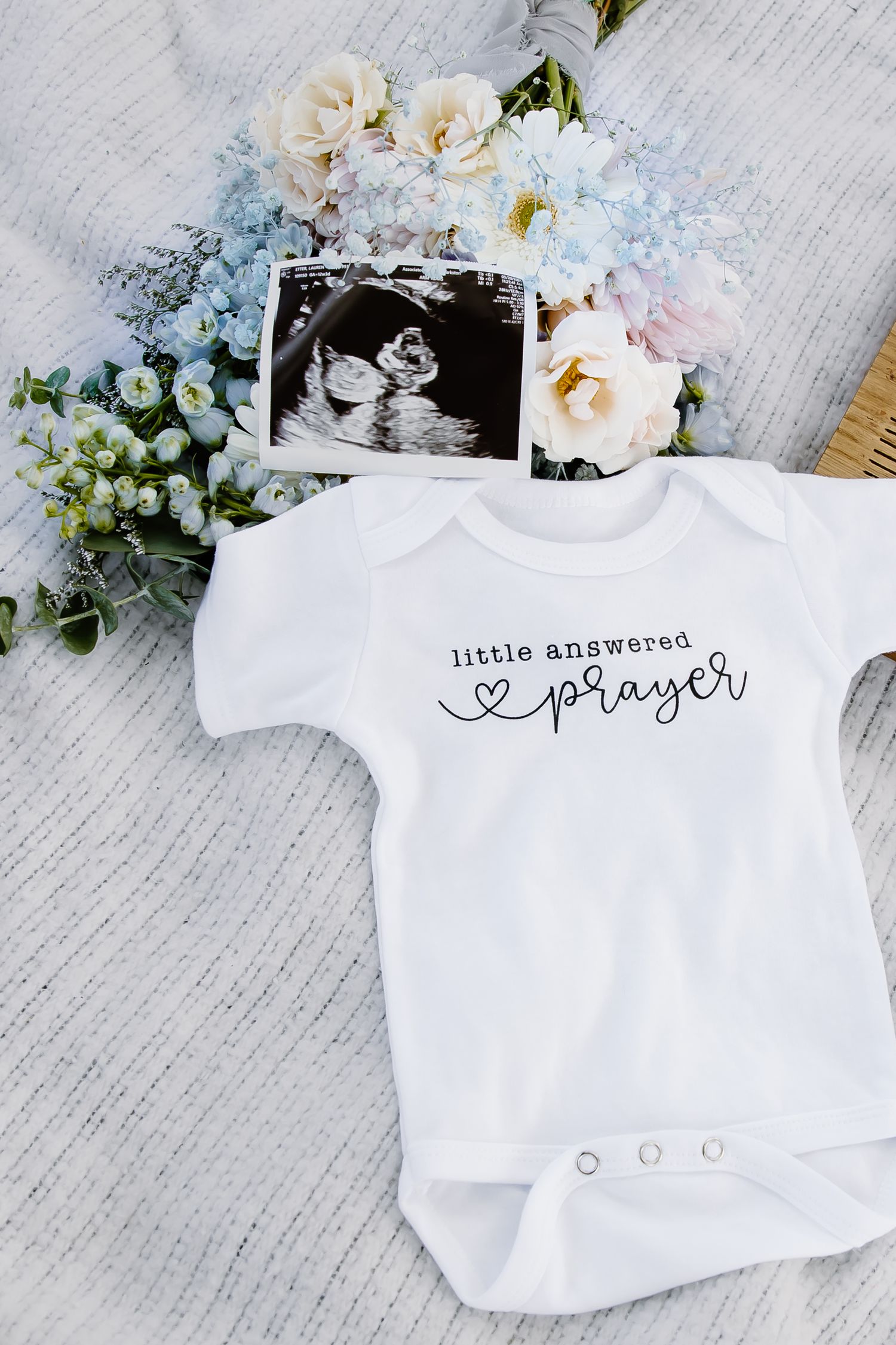Baby onesies with 'little answered prayer' text laid out with flowers and ultrasound photos.