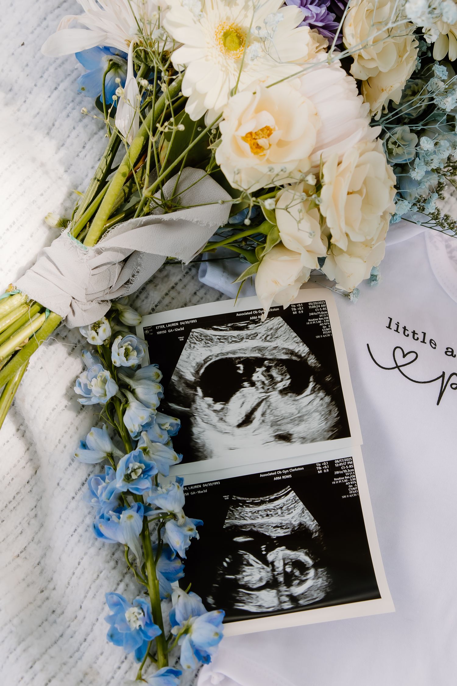 Elegant floral arrangement with ultrasound photos and personalized baby onesie on white fabric.