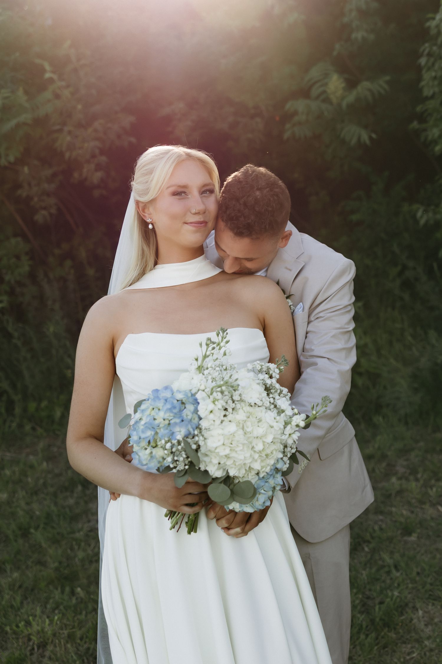 Bride in white strapless gown holds blue and white bouquet as groom in tan suit nuzzles her neck outdoors.