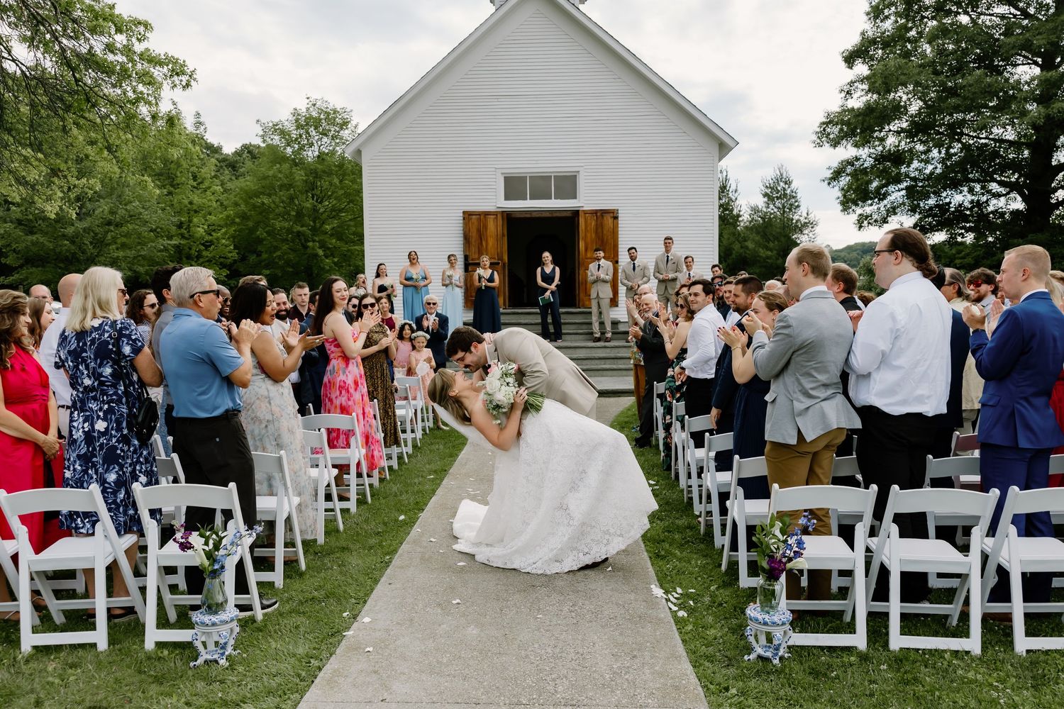 Bride walking down outdoor wedding aisle in white gown past seated guests toward small white chapel.