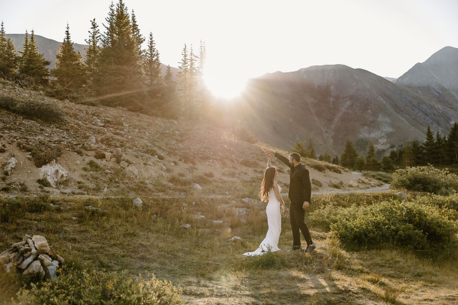 Couple in wedding attire walking mountain path bathed in warm sunset light with peaks in background.
