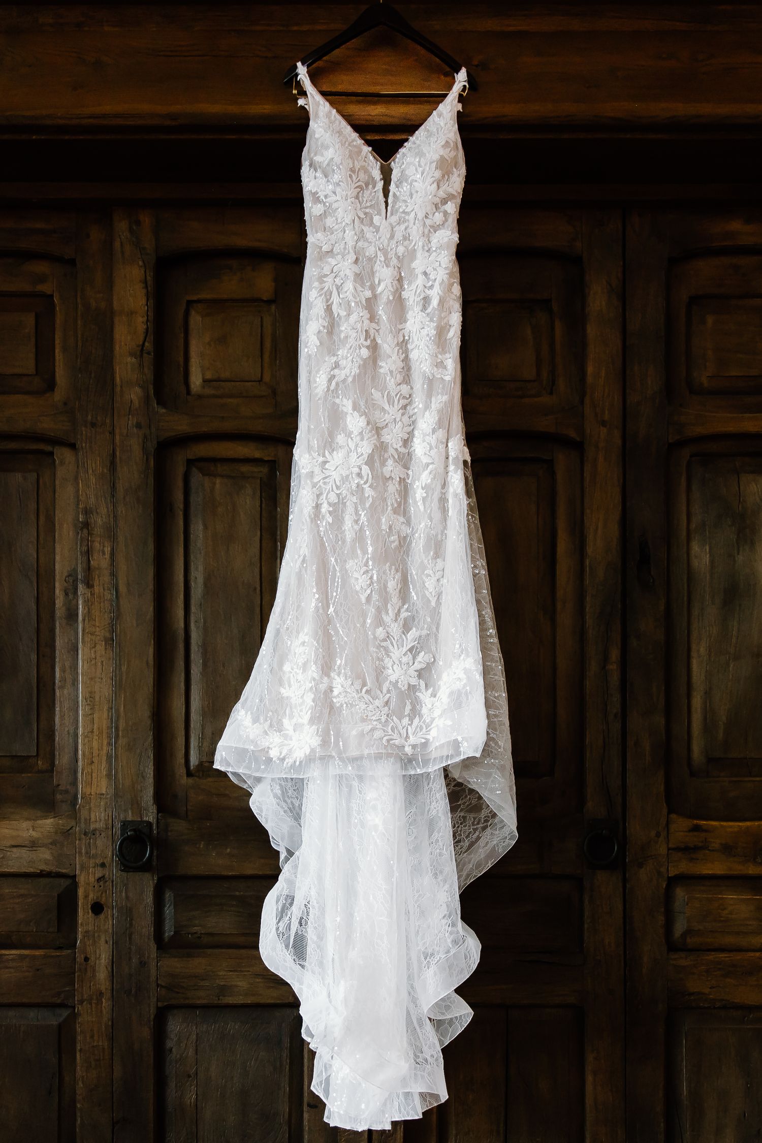 Delicate lace wedding dress hanging against dark wooden paneled wall.