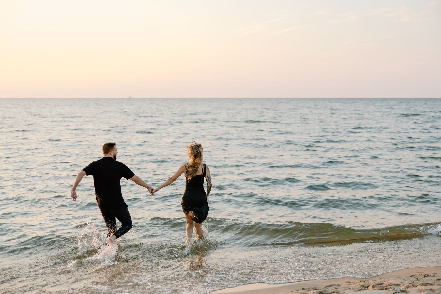 Couple splashing in shallow ocean waves at sunset while holding hands.