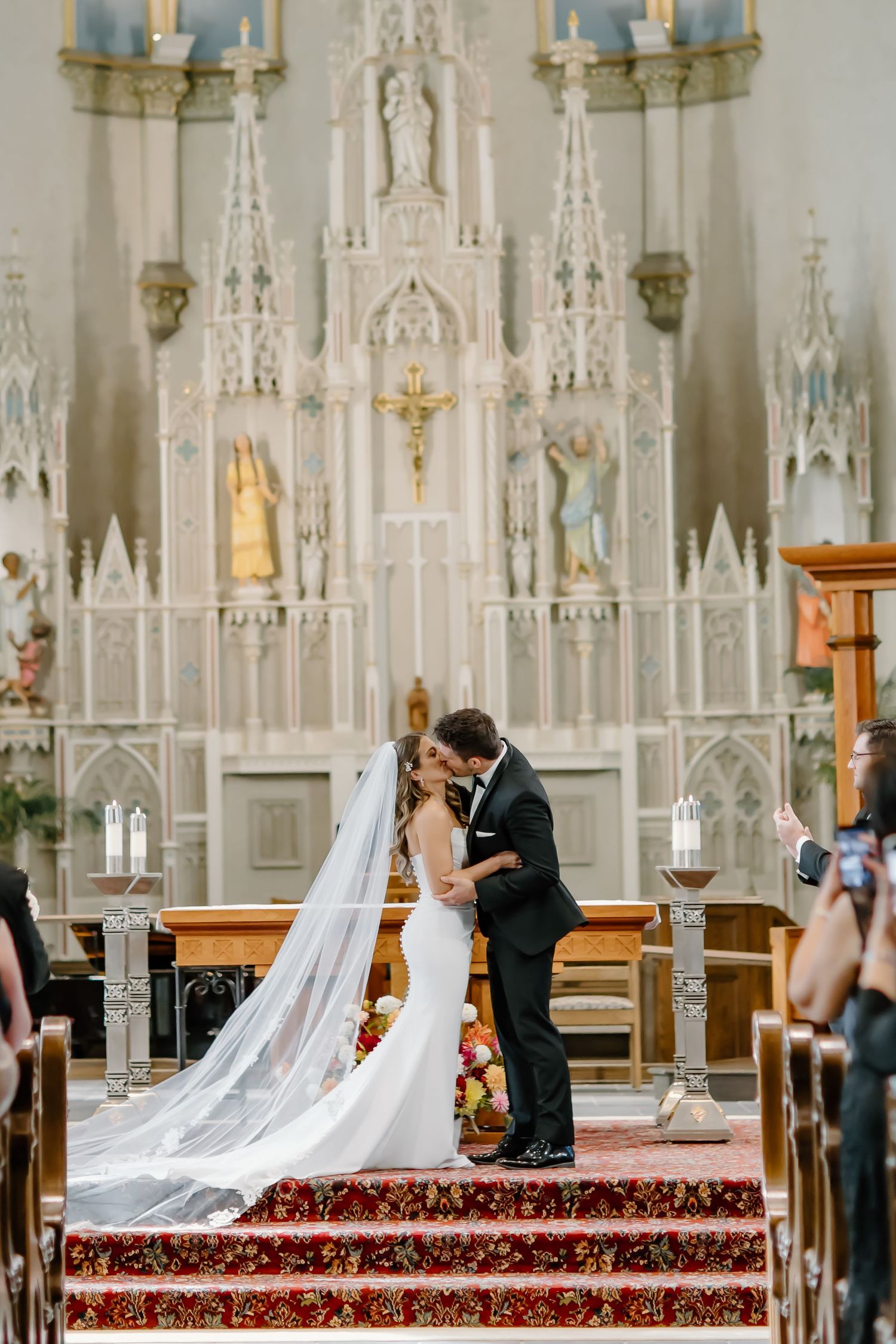 Wedding ceremony at ornate white Gothic altar with red carpeted steps and candlesticks.