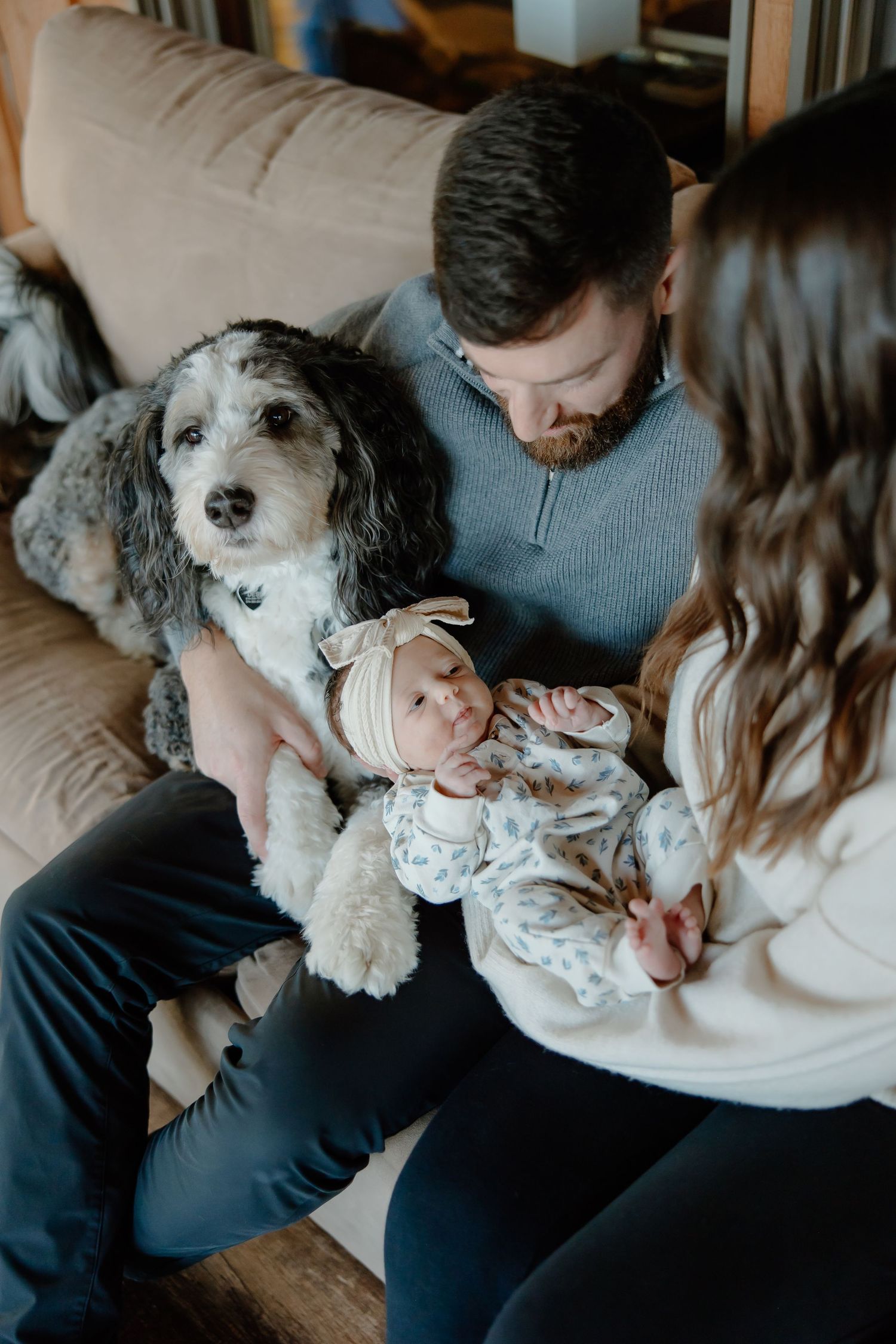 A family relaxes on the couch with their fluffy dog and newborn baby in a heartwarming scene.