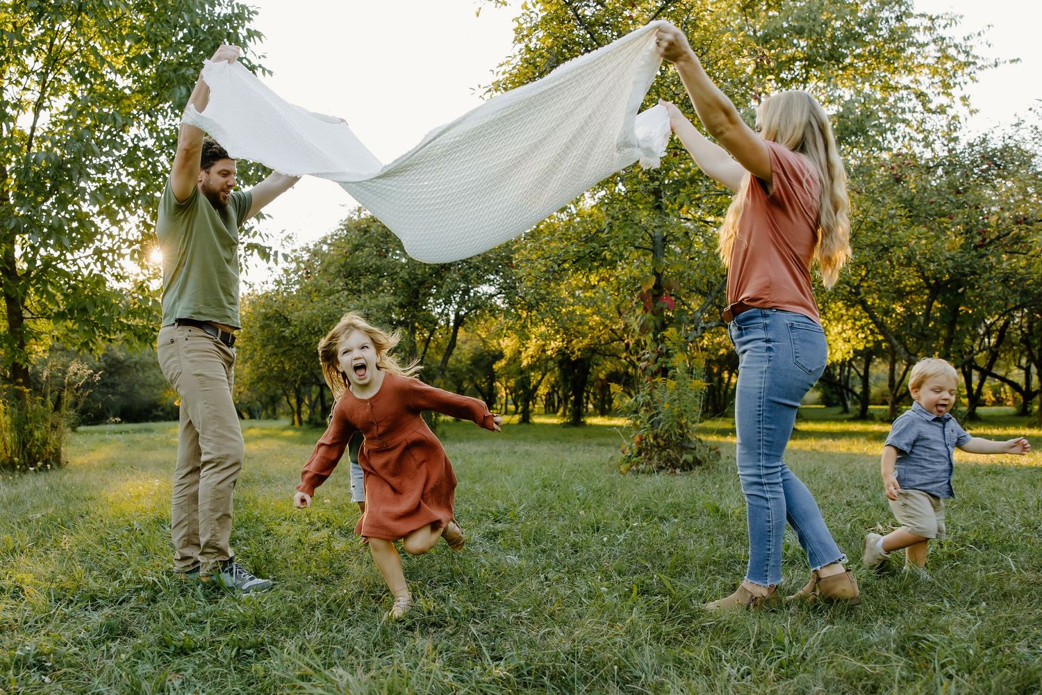 Family playing under a white sheet in an orchard during golden hour.