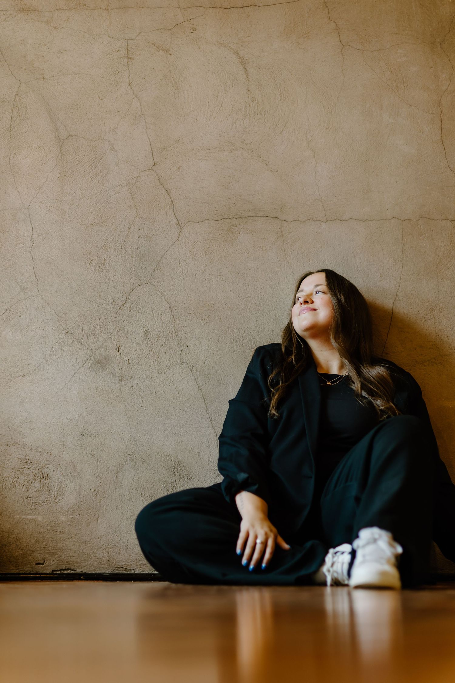 Person in black outfit sits on wooden floor against textured wall with eyes closed and relaxed expression.