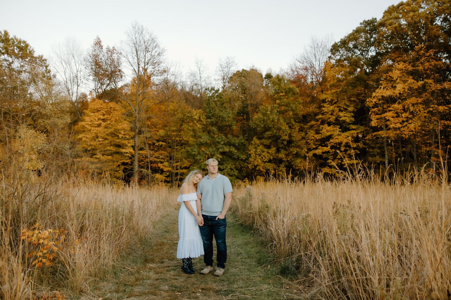 Couple standing in tall grass with autumn foliage behind them during golden hour.