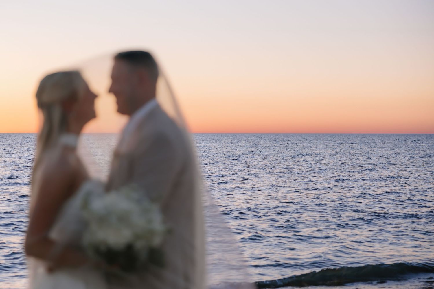 Bride and groom share a romantic moment at sunset by the ocean, with a white veil softly framing the couple.