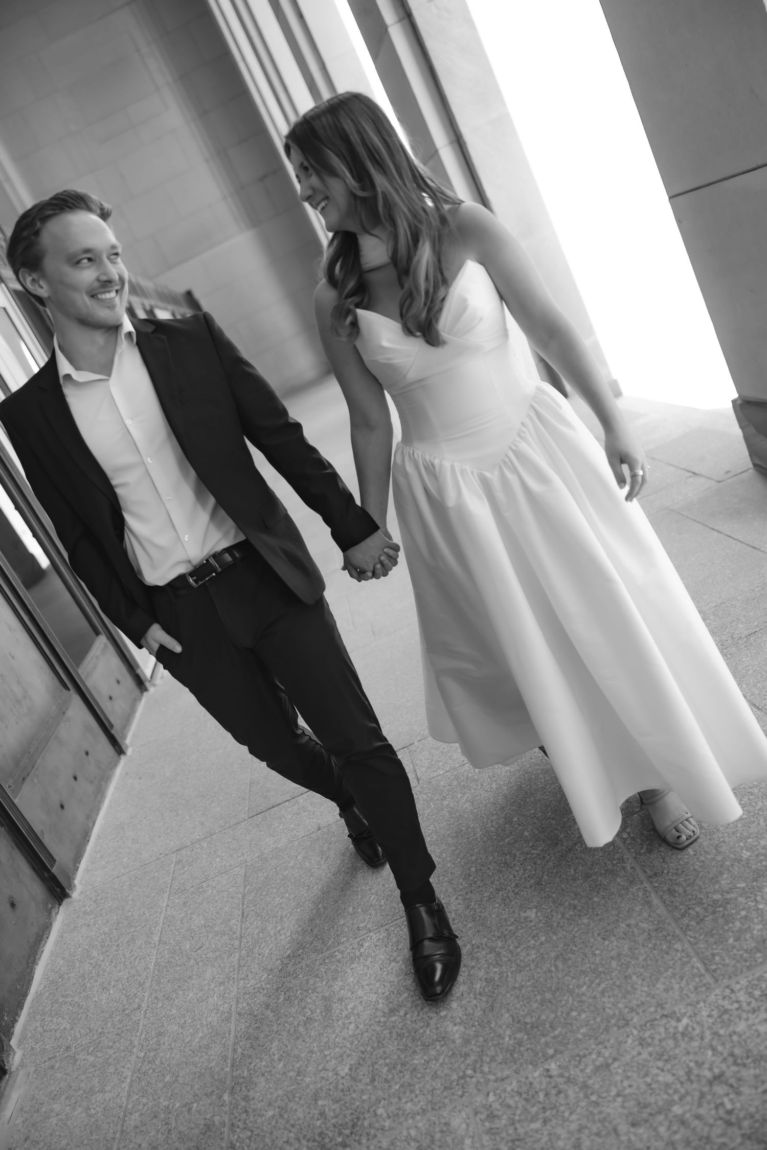 Black and white photo of a smiling couple in wedding attire holding hands and walking together outside a building.