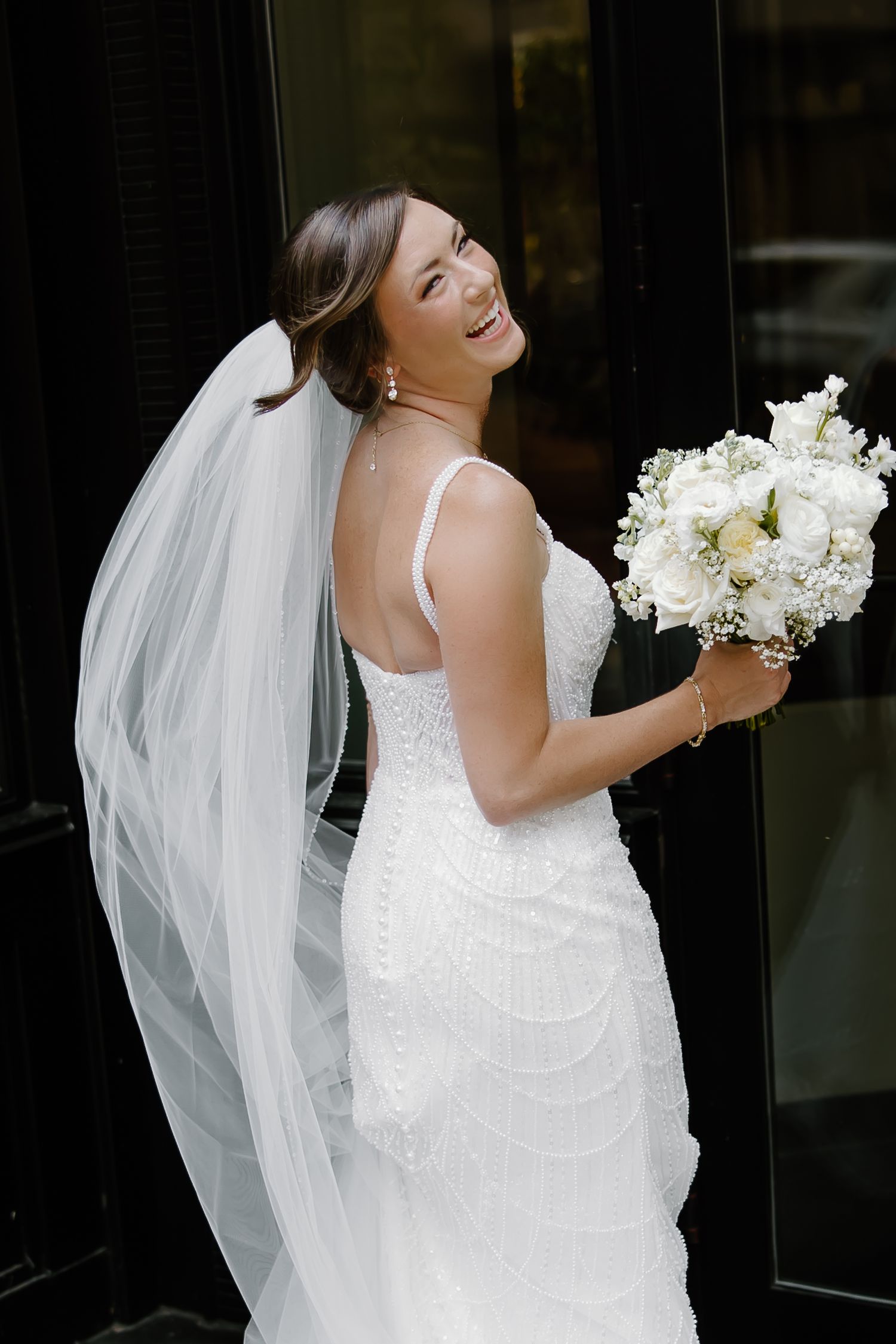 Joyful bride in fitted lace gown holds white bouquet while posing against dark background.