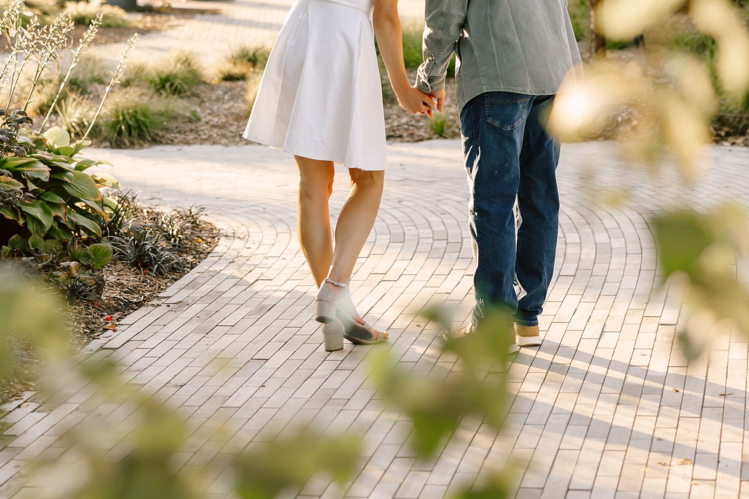 Couple walking on sunlit brick pathway through garden setting.