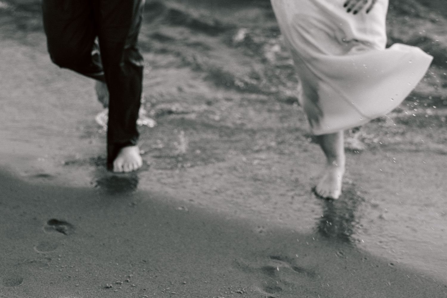 Black and white photo of couple's feet walking along wet beach sand.