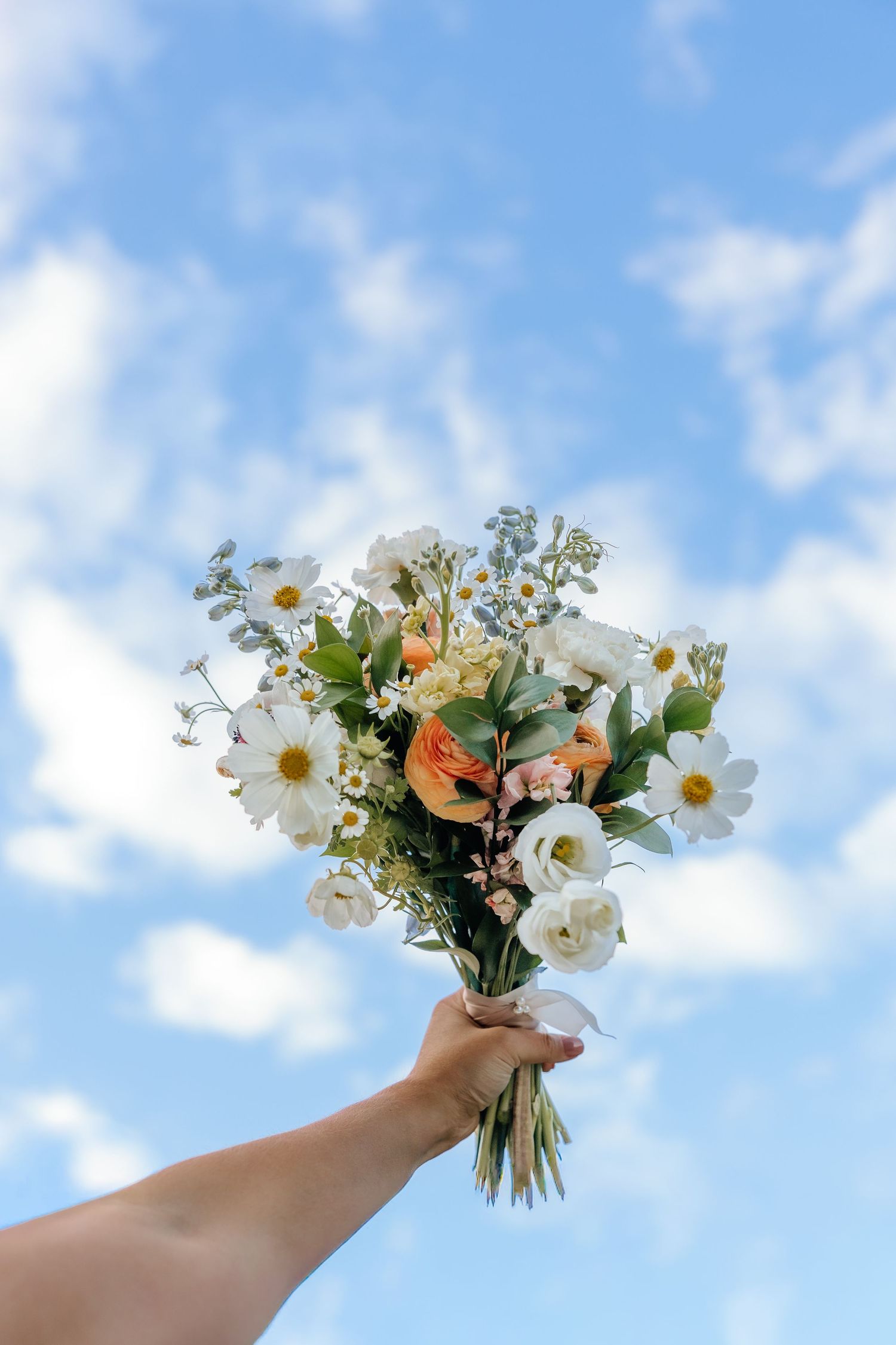Hand holding a delicate bouquet of white daisies and peach flowers against a bright blue sky with clouds.