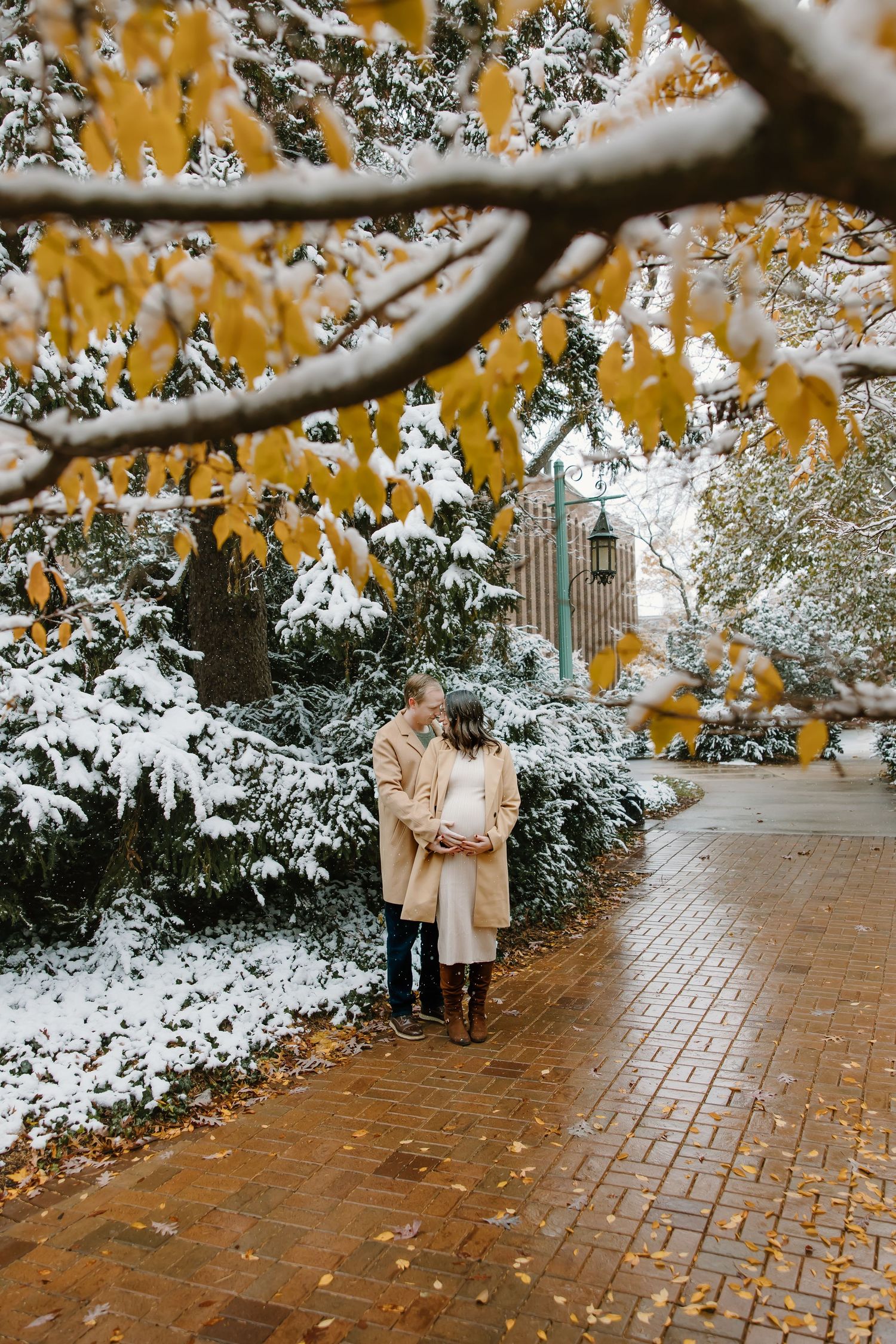 Couple in tan coats walking along snowy brick path under golden autumn leaves and gothic architecture.