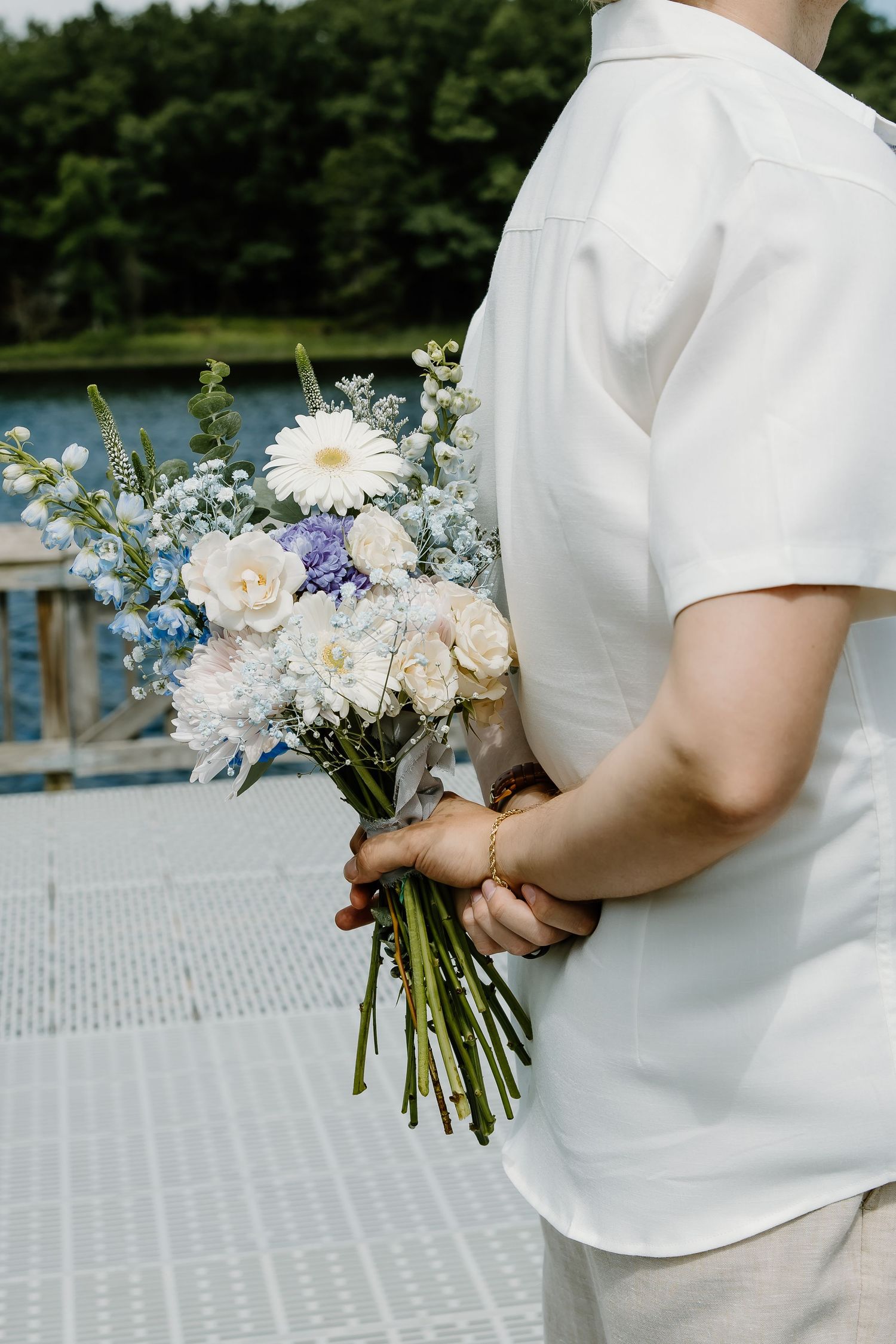 Beautiful wedding bouquet with white daisies, roses, and blue delphinium flowers against a waterfront backdrop.