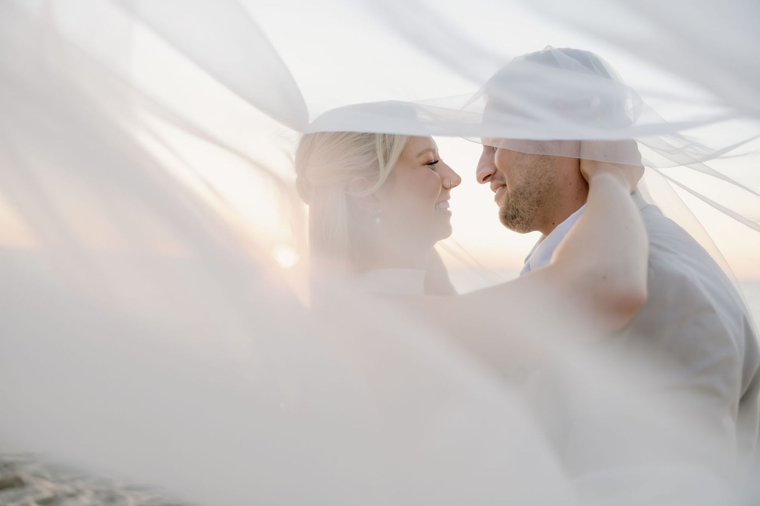 Romantic couple sharing an intimate moment under a flowing white veil at a beach wedding.