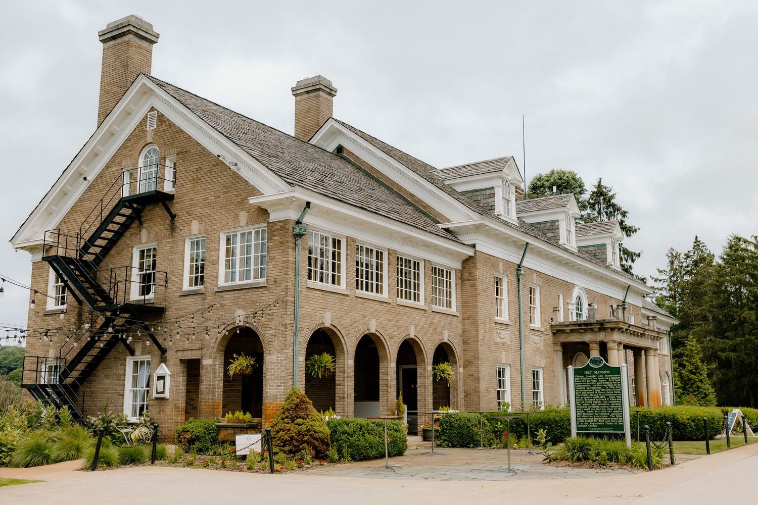 Historic brick manor house with arched entryway, fire escape, and lush gardens on overcast day.