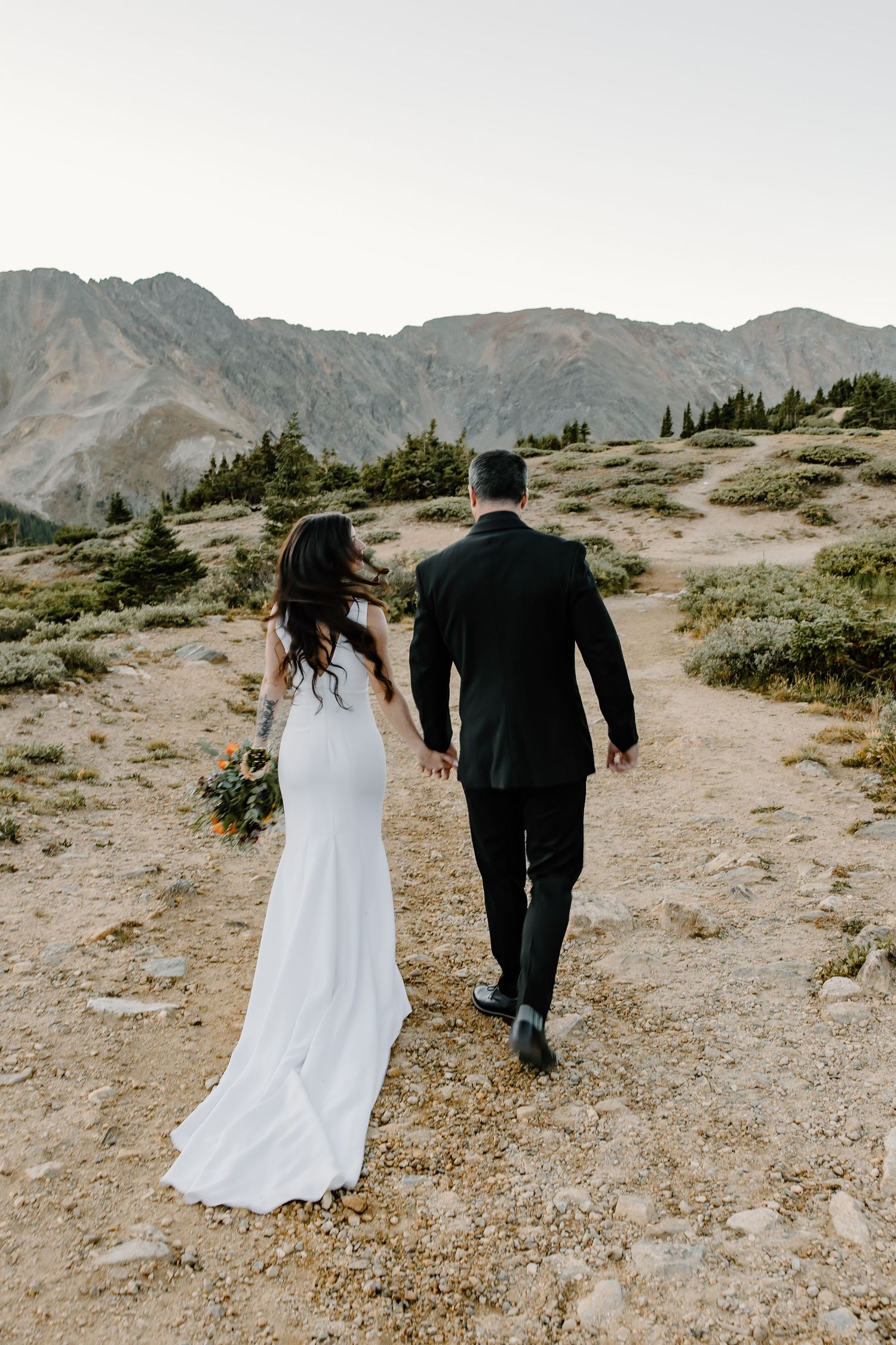 A couple walks away from camera on mountain path wearing wedding attire against dramatic alpine backdrop.