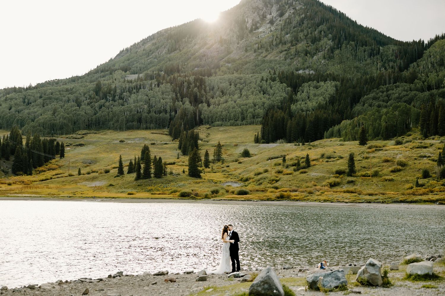Silhouetted wedding couple by mountain lake with sunburst through hills.
