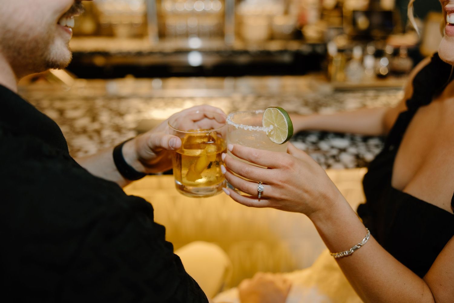 Two people toasting with drinks at an illuminated bar counter.