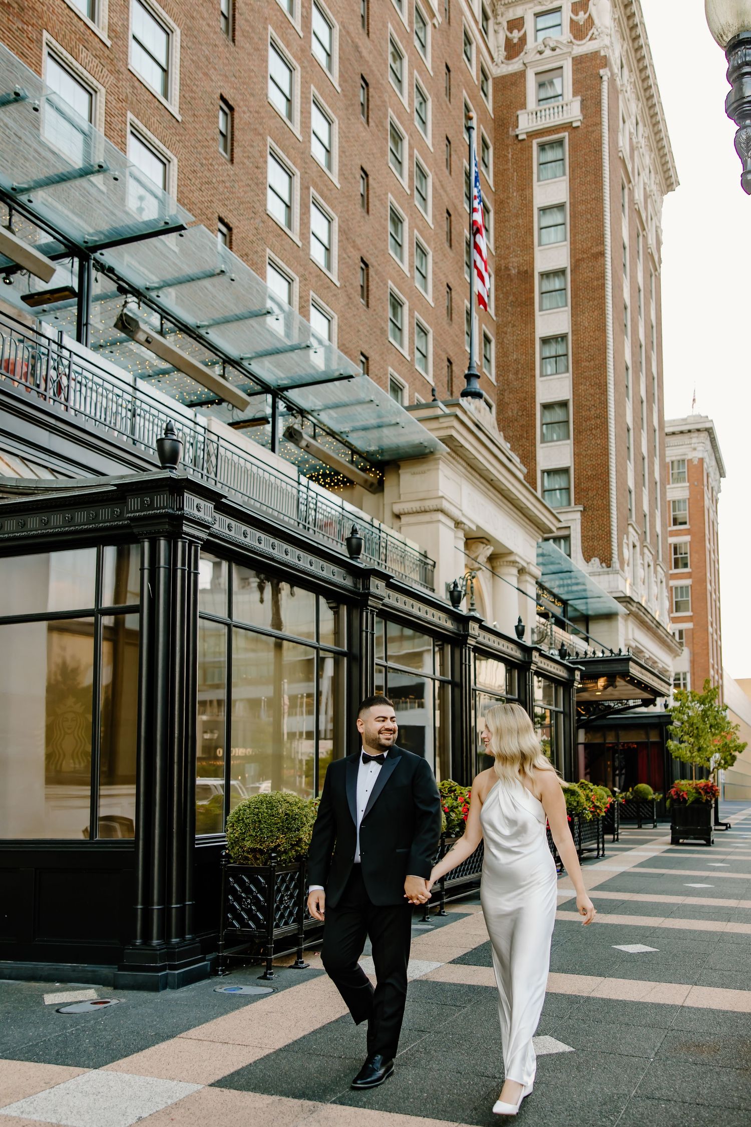 Elegant couple walks hand in hand outside historic brick building with glass awning.