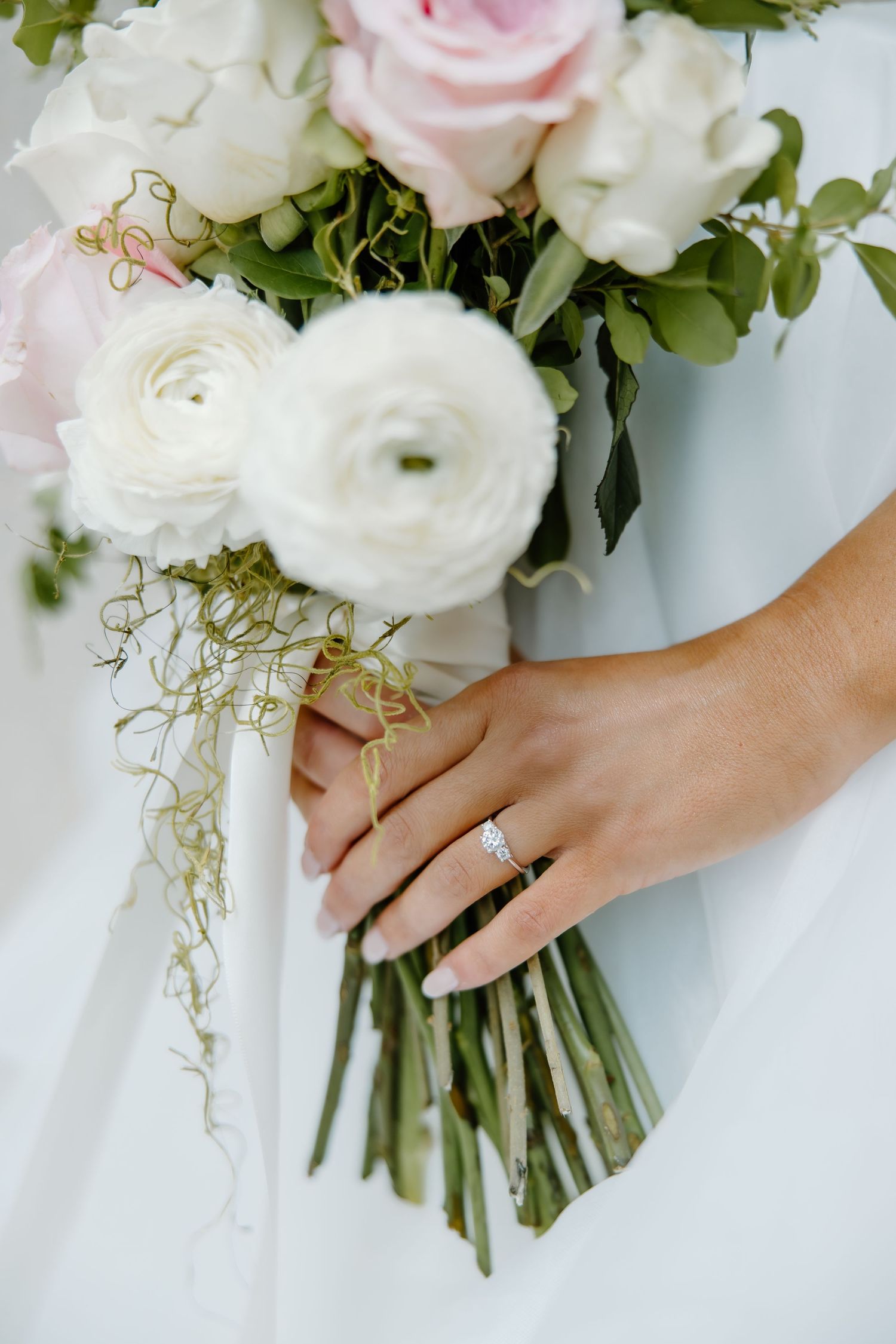 Close-up of a bridal bouquet with white ranunculus and pink roses, showing an engagement ring on the bride's hand.
